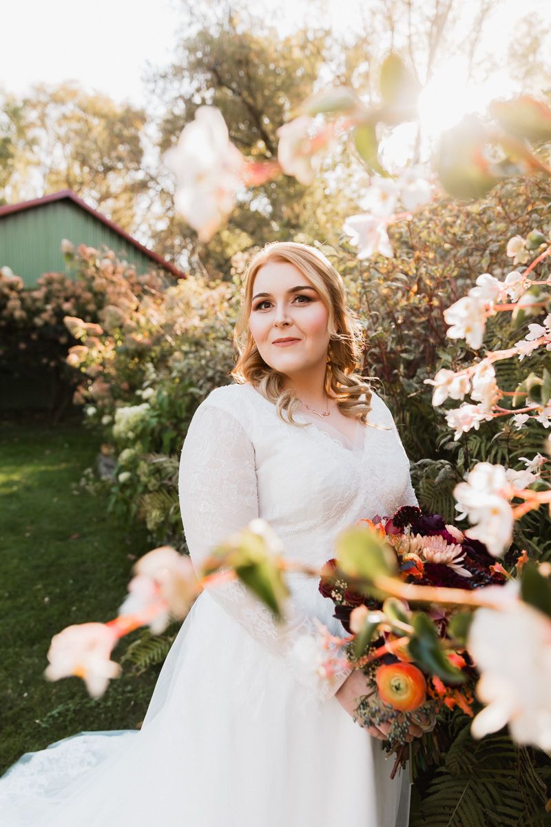 A bride in a white lace dress poses among blooming flowers during a romantic golden hour photoshoot.