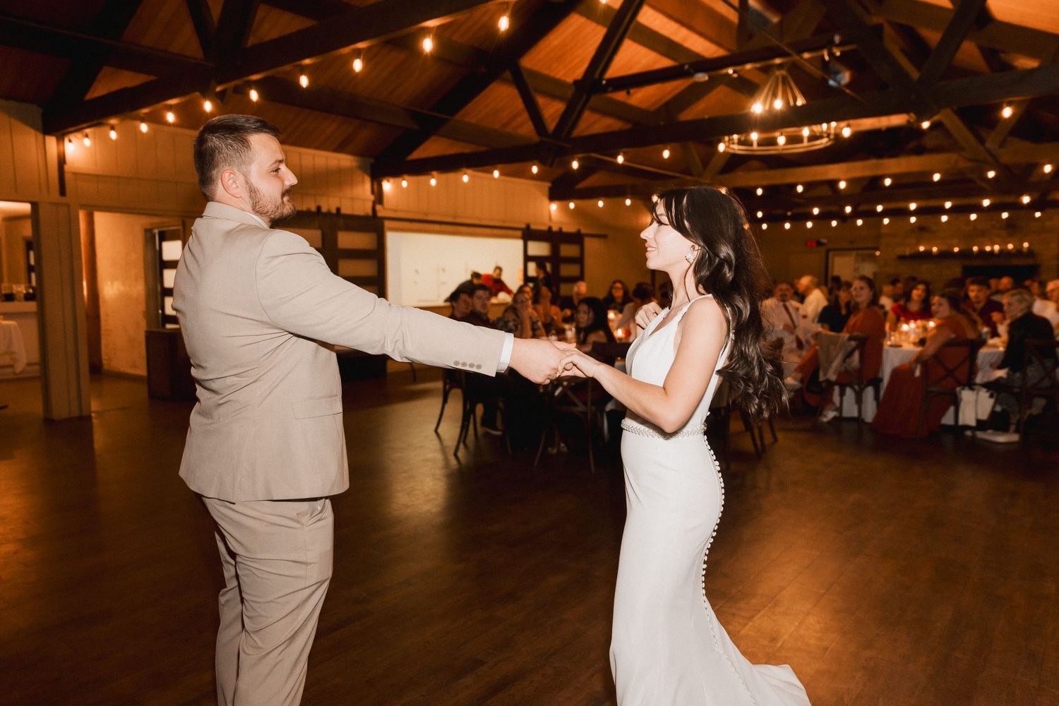 A couple dances together in a rustic reception venue with warm lighting and exposed wooden beams overhead.