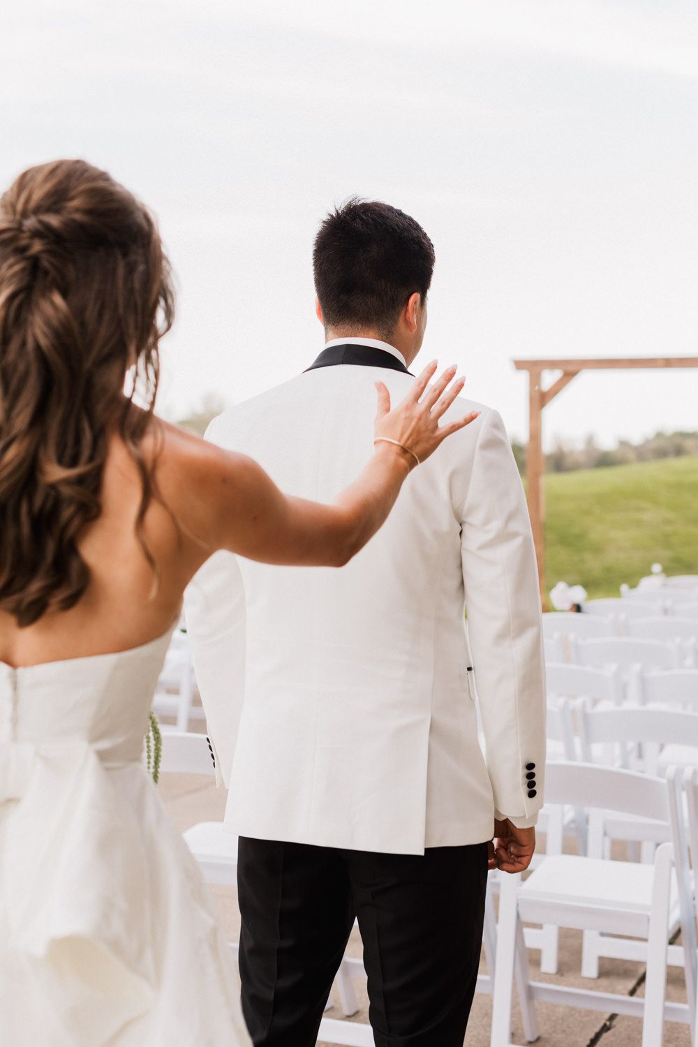 A couple in wedding attire holds hands and waves while walking away from the camera in an outdoor venue setting.