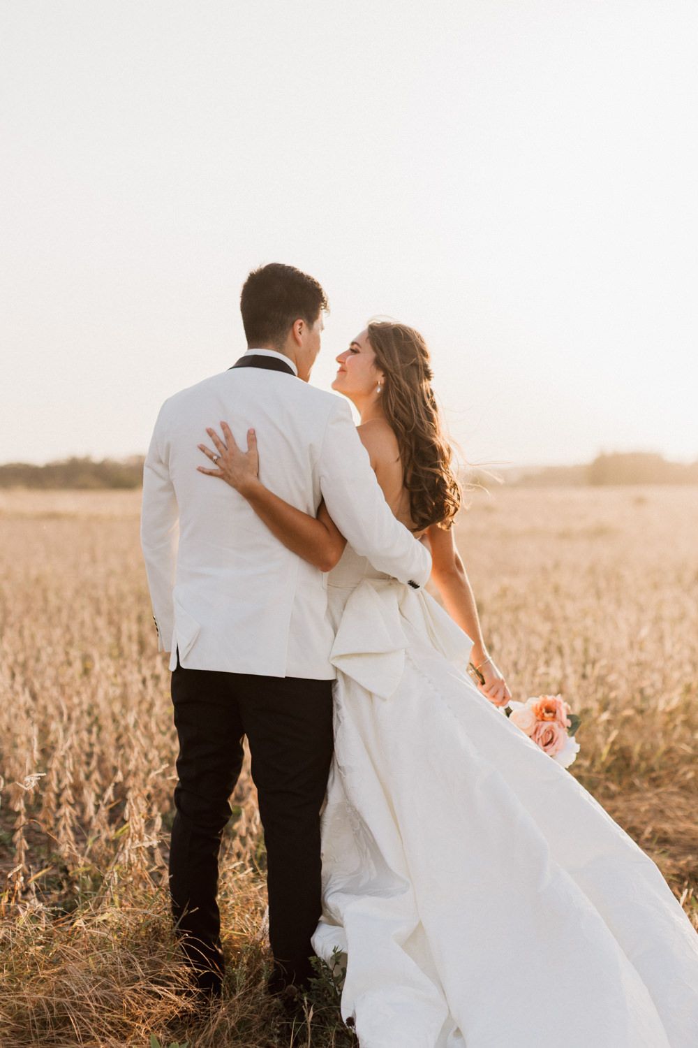 Newlywed couple embracing in a golden wheat field at sunset with flowing white wedding dress.