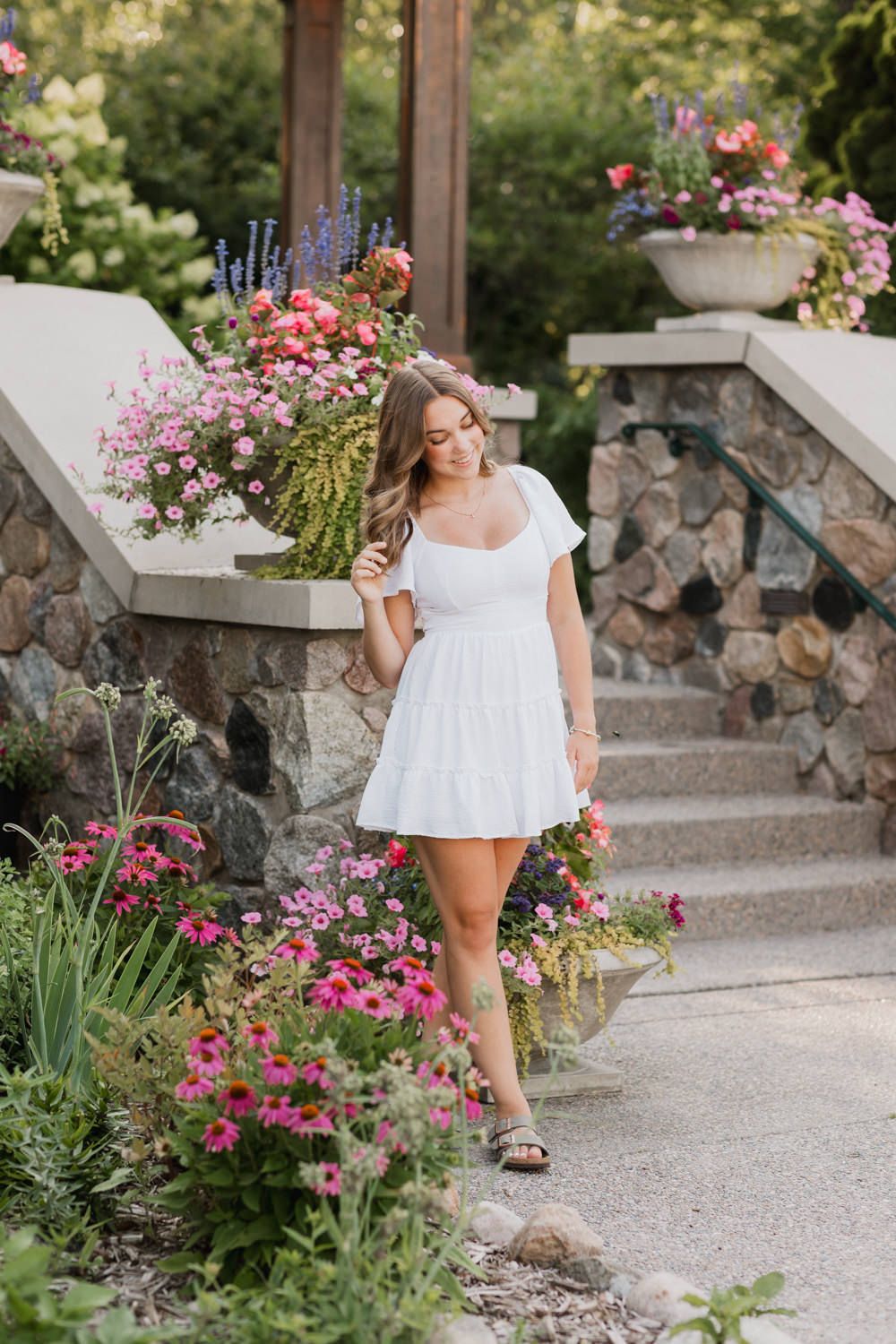 A person in a white summer dress poses gracefully among pink flowers and stone steps in a garden setting.