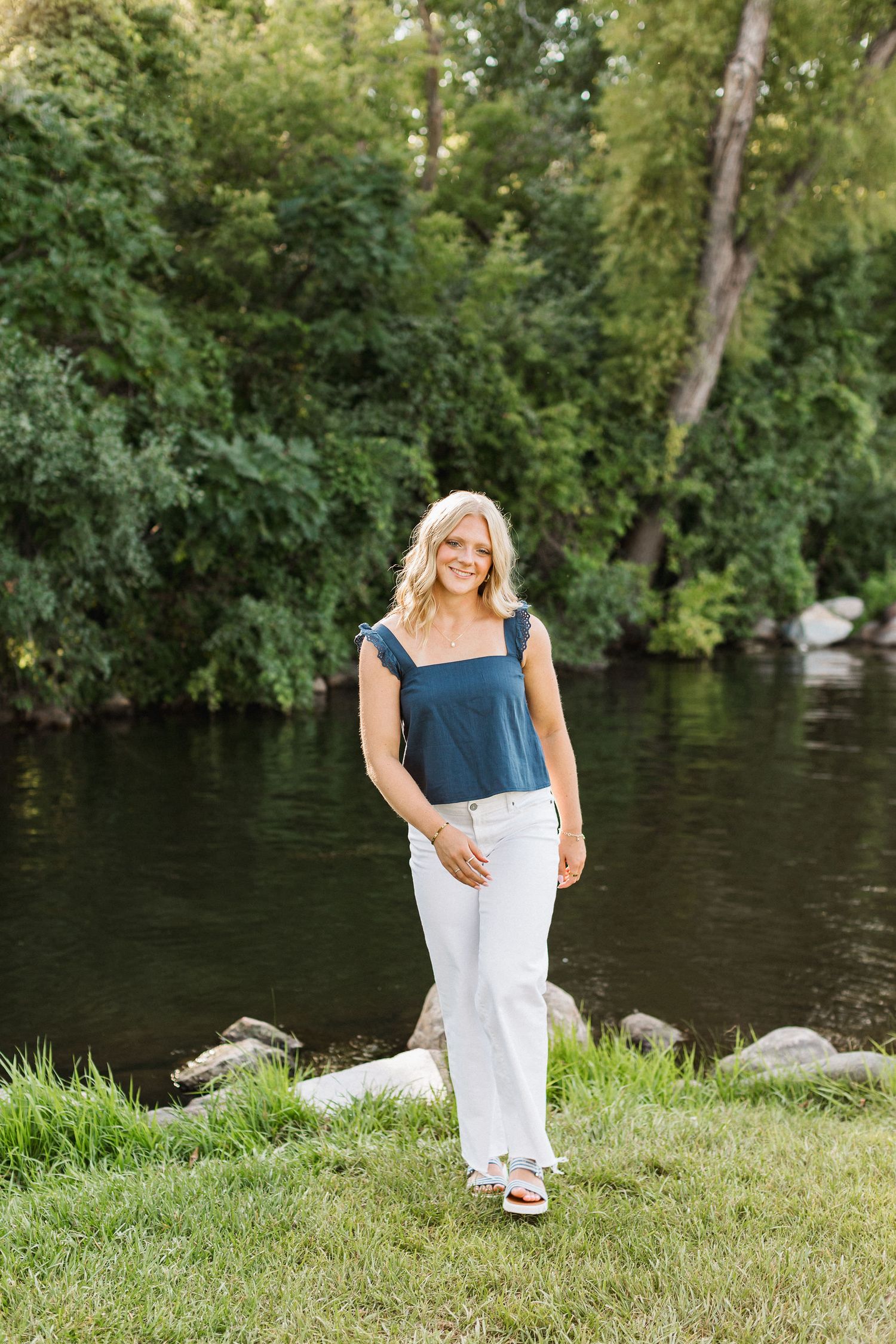 A person in a blue top and white skirt sits by a peaceful riverbank surrounded by lush green trees and rocks.
