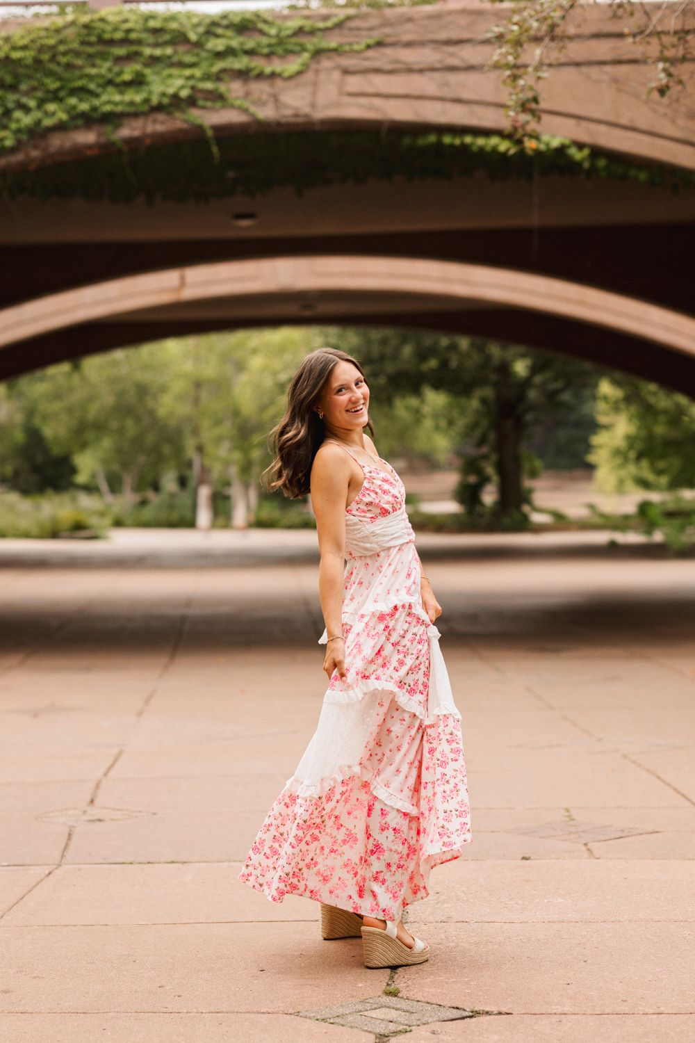 A person in a pink floral maxi dress twirls under a stone bridge on a sunny summer day.