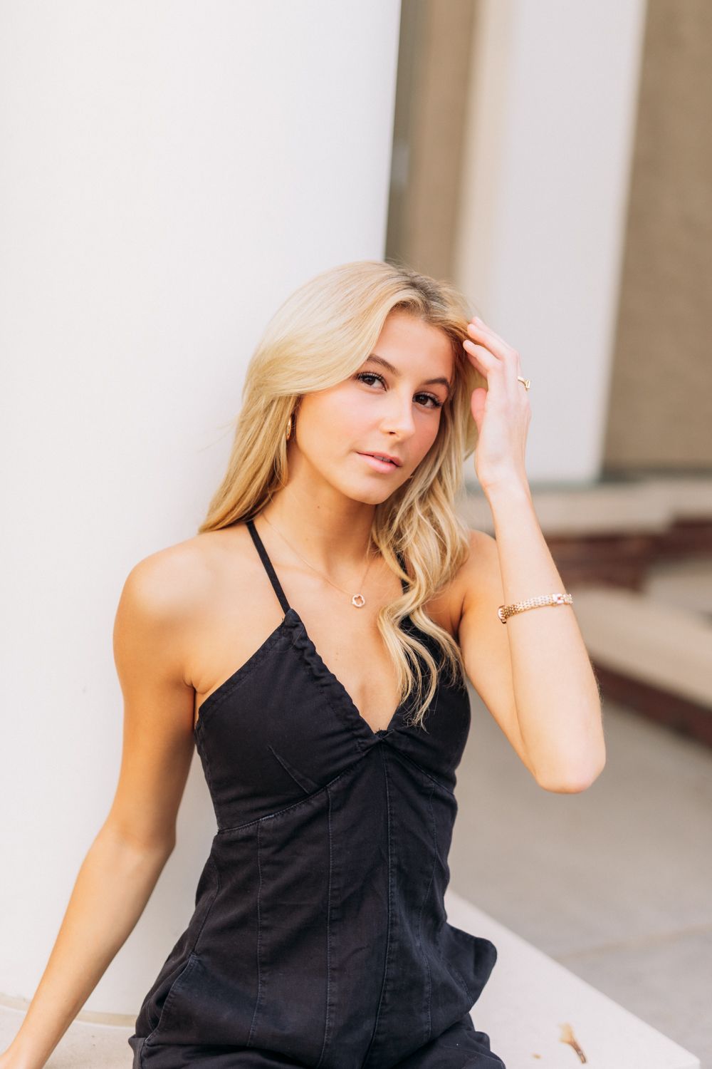 Blonde woman in black v-neck dress poses outdoors against white wall background.