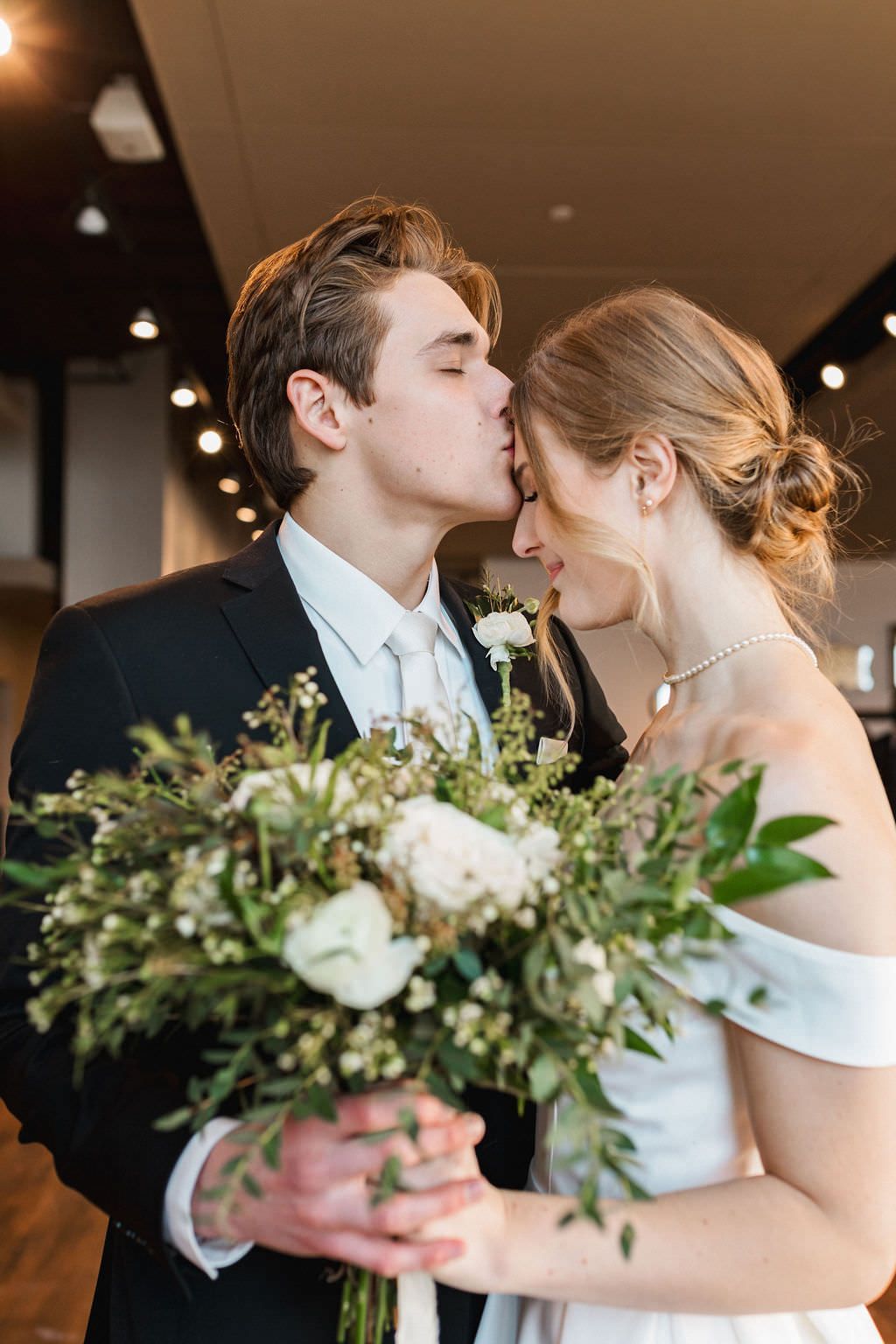 A romantic wedding couple shares an intimate kiss while holding a white and green bouquet in an indoor venue.