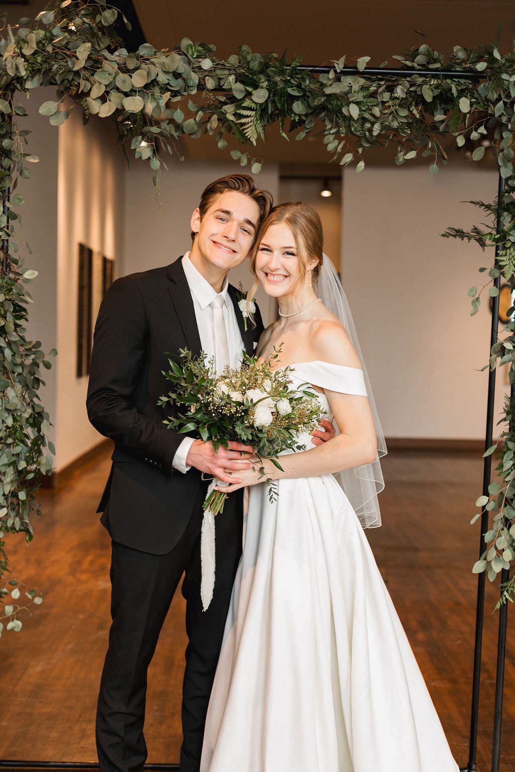 Romantic moment captured under floral arch with brick walls and hardwood floors visible in the background.
