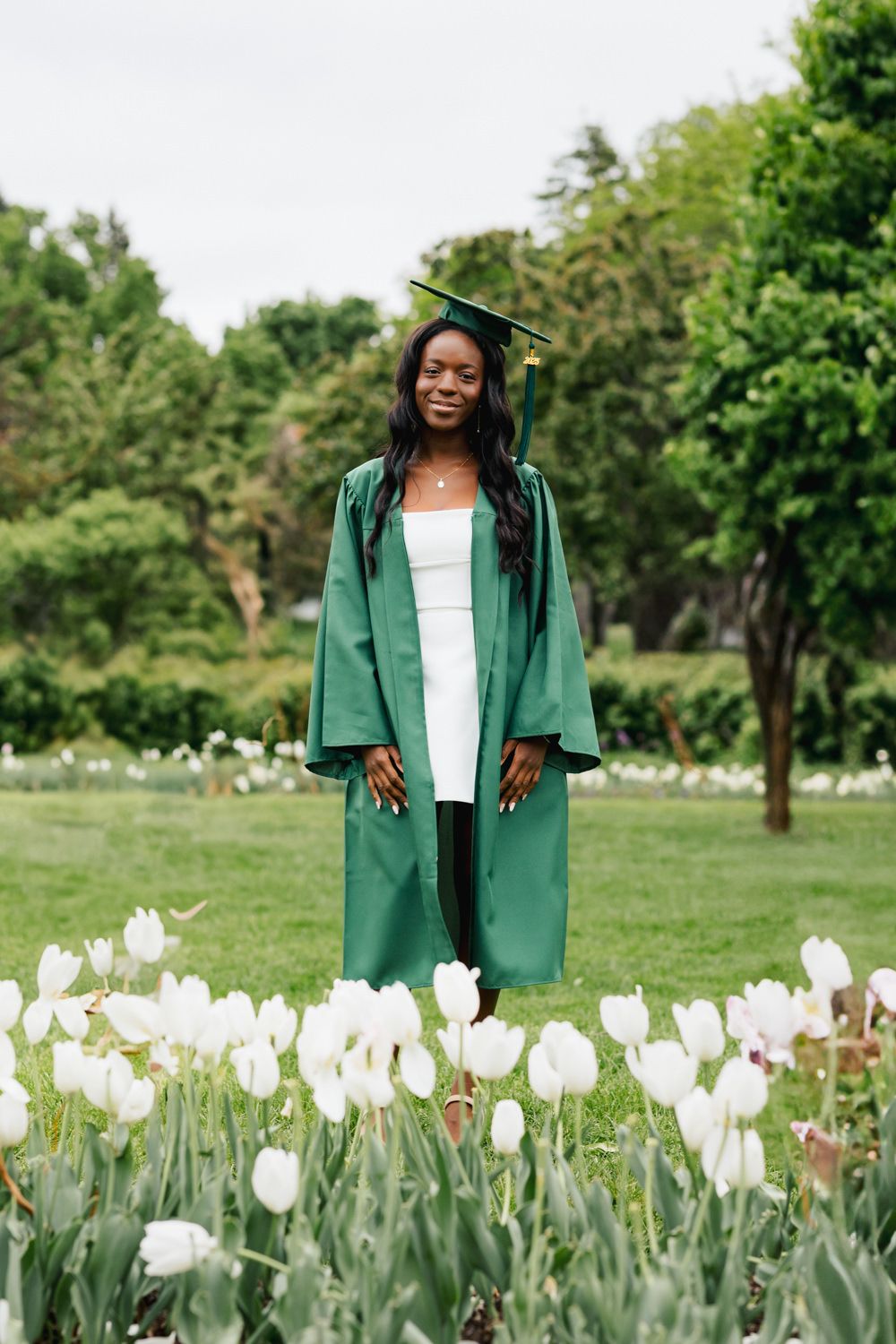 Graduate in emerald regalia stands among blooming white tulips in garden setting.