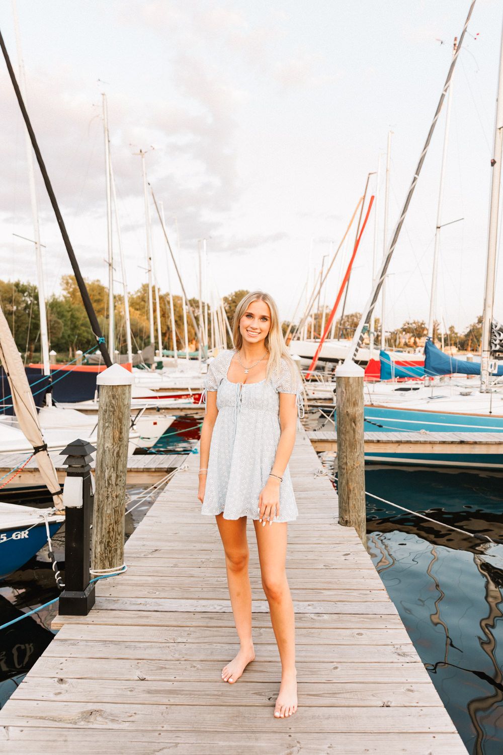 Model in white dress poses barefoot on marina dock with sailboats in background.
