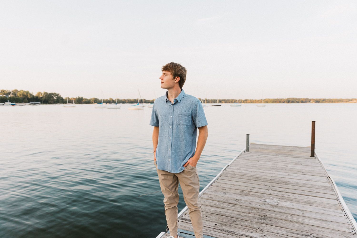 Person in blue shirt stands on wooden dock overlooking serene lake at sunset.