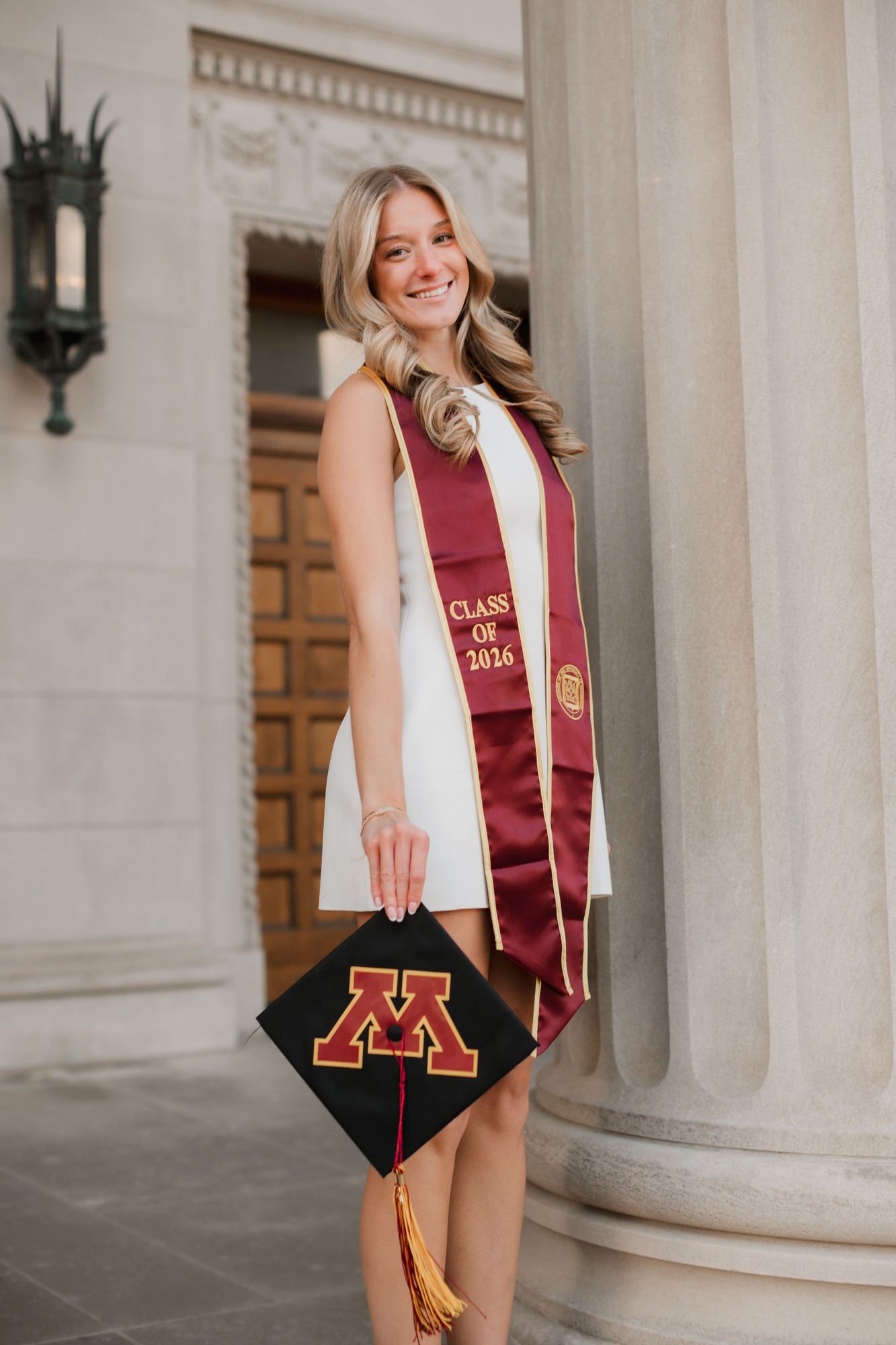 University of Minnesota graduate in white dress holding maroon Class of 2026 stole and decorated graduation cap.