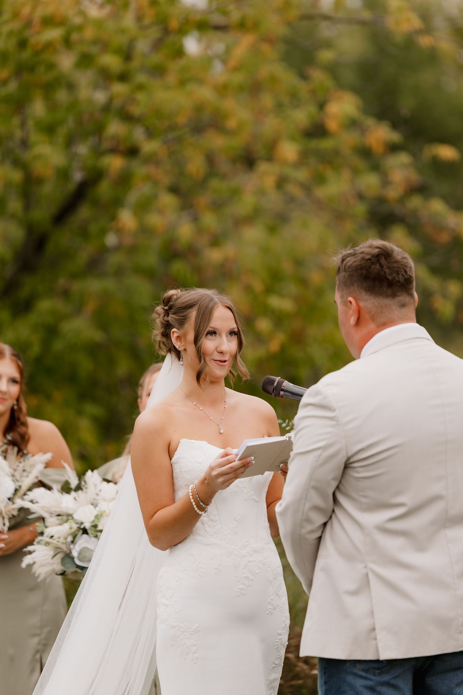 Bride reads vows to groom outdoors during fall wedding ceremony with colorful foliage backdrop.