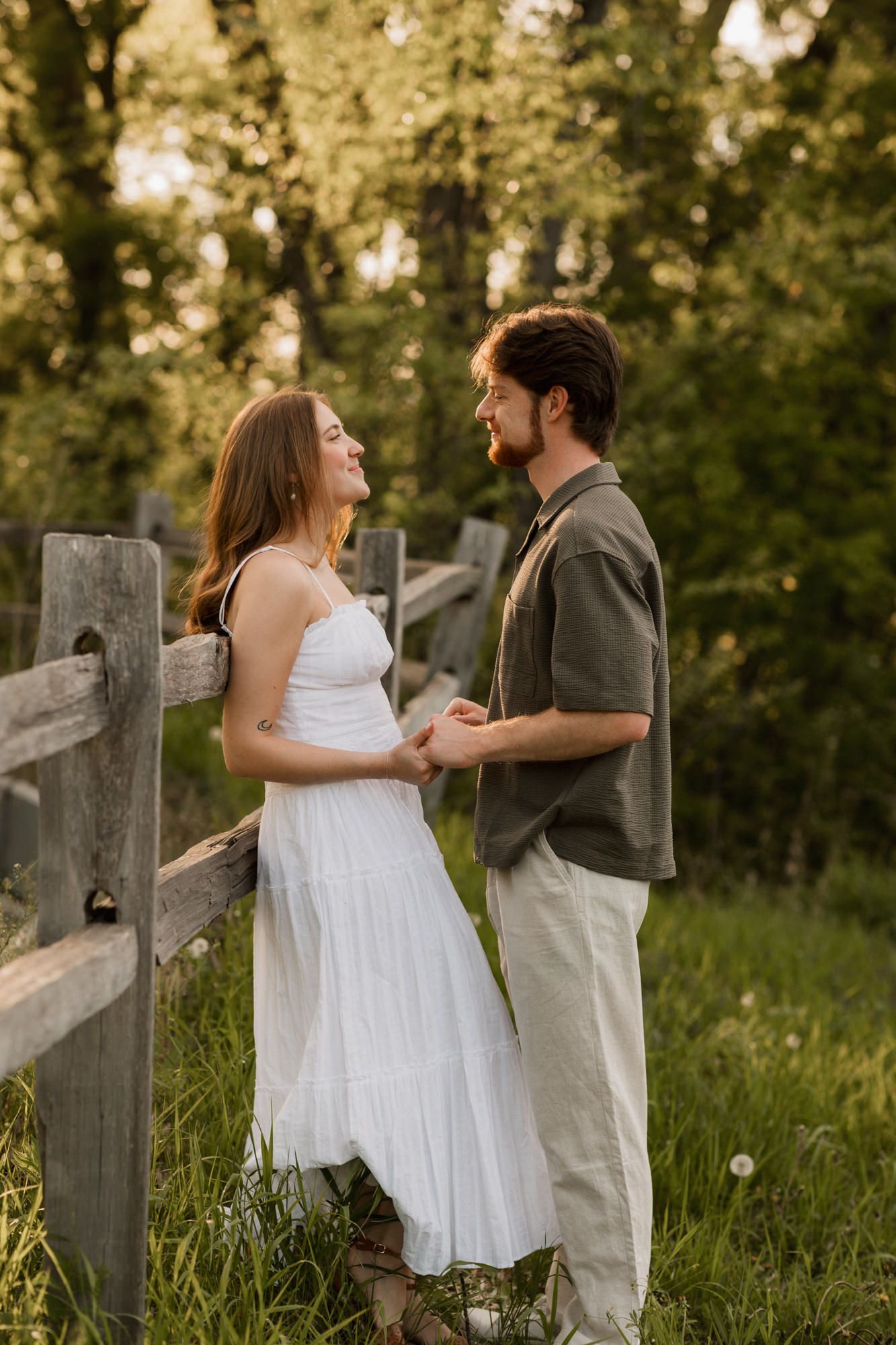 Couple holding hands and smiling at each other by a wooden fence surrounded by lush green trees in golden hour light.