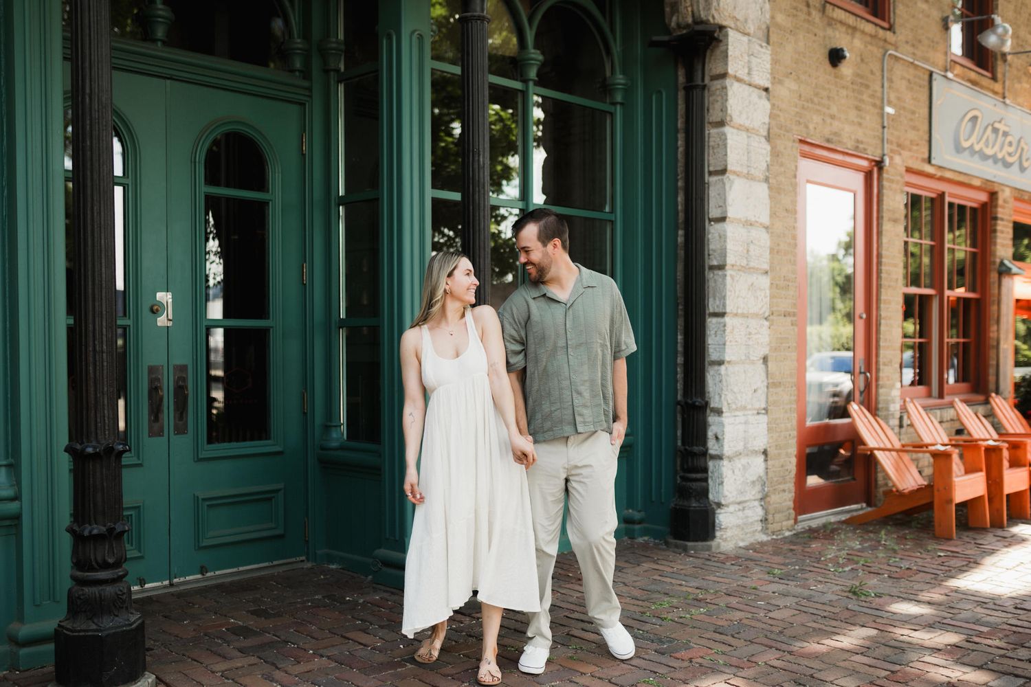 Couple holding hands smiling at each other outside a green storefront in a historic brick district.