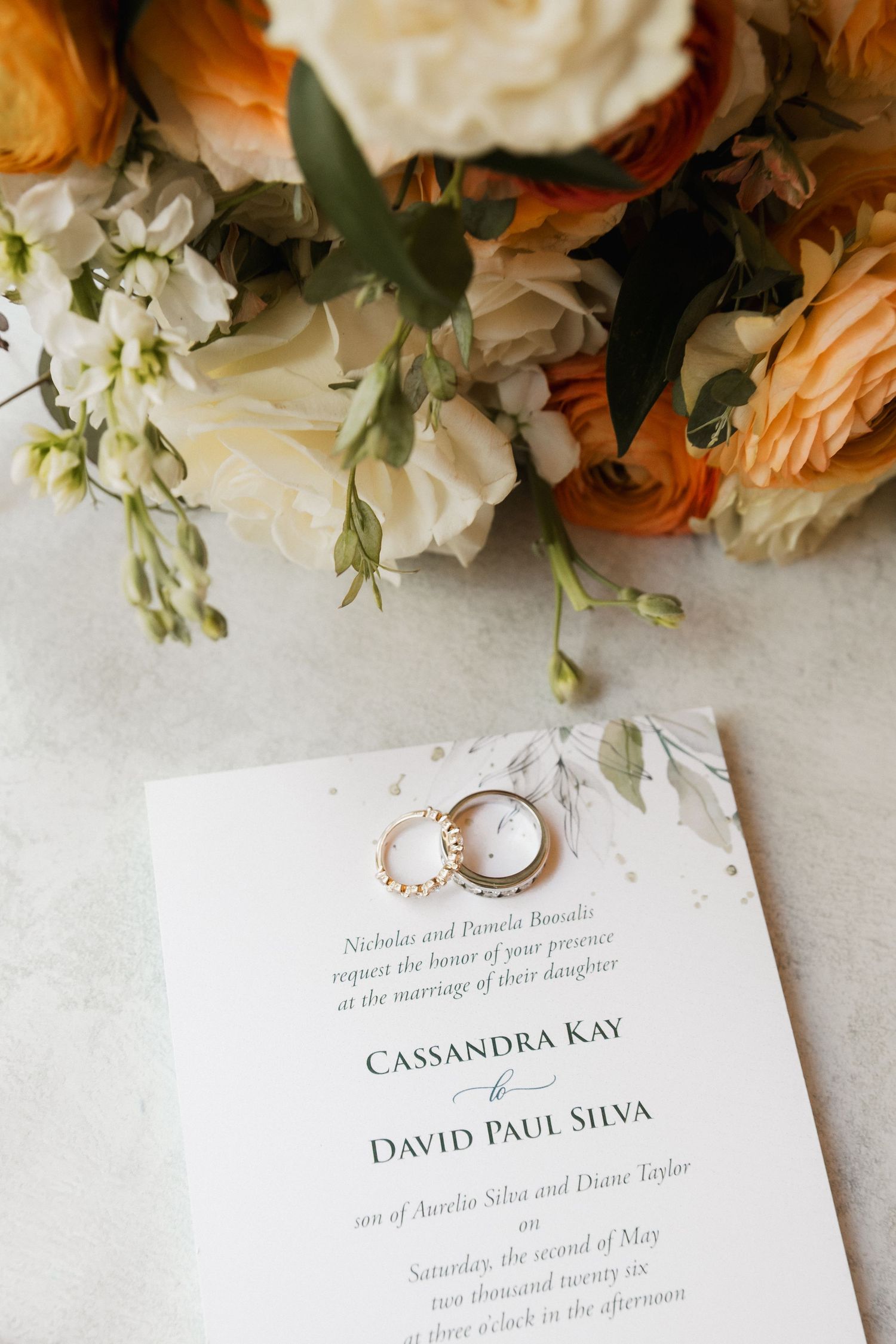 Wedding invitation for Cassandra Kay and David Paul Silva with gold rings resting on it, surrounded by orange and white floral bouquet.