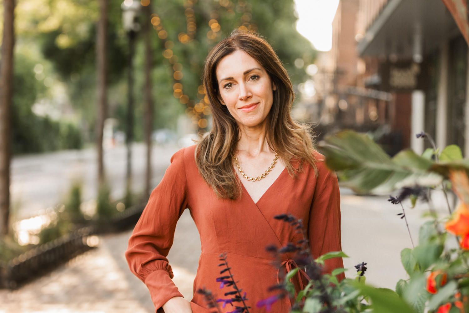 Woman in rust-colored dress smiling outdoors on a tree-lined urban street surrounded by flowers.