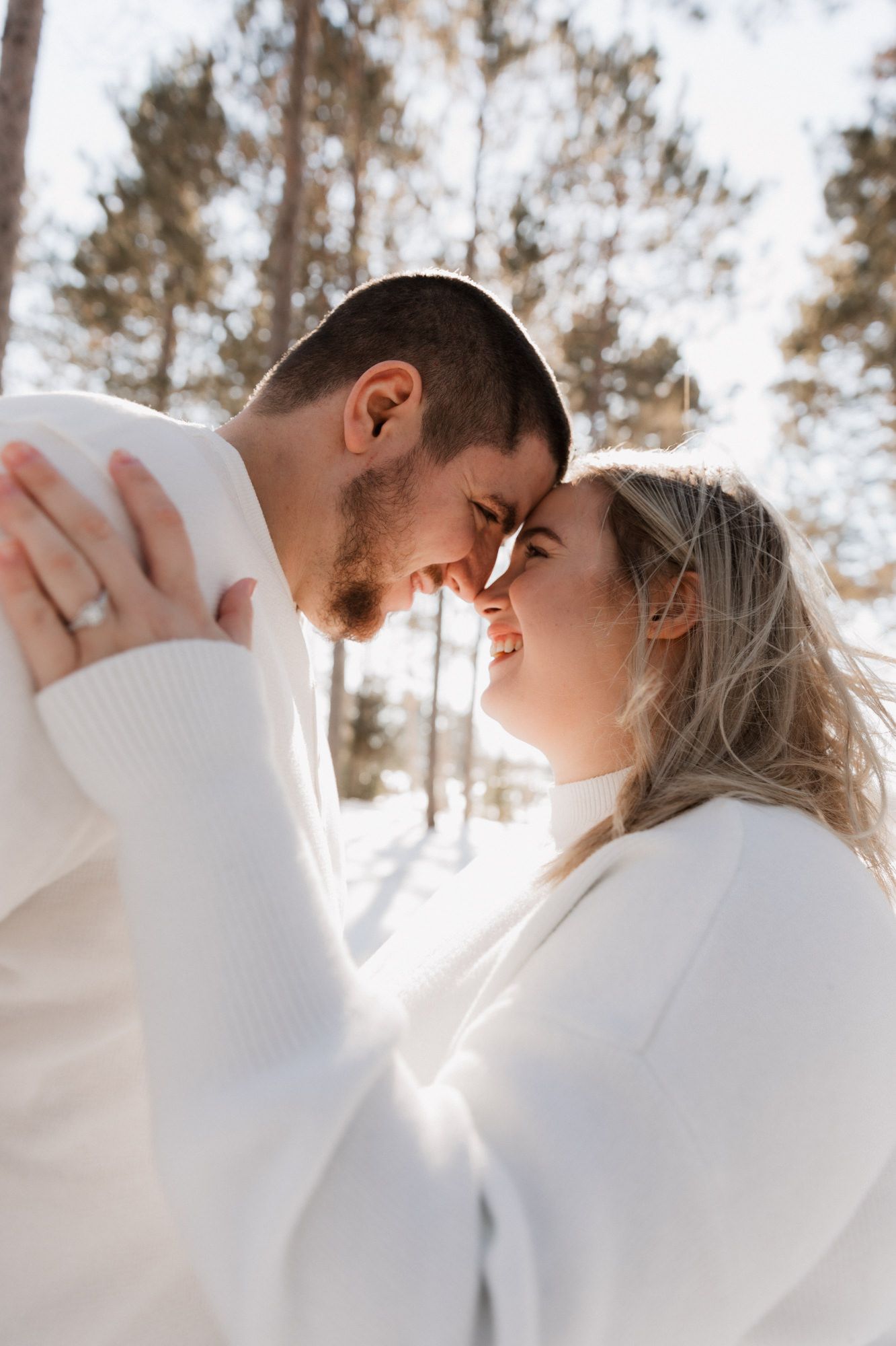 Couple in white sweaters touching foreheads and smiling in a snowy winter forest setting.