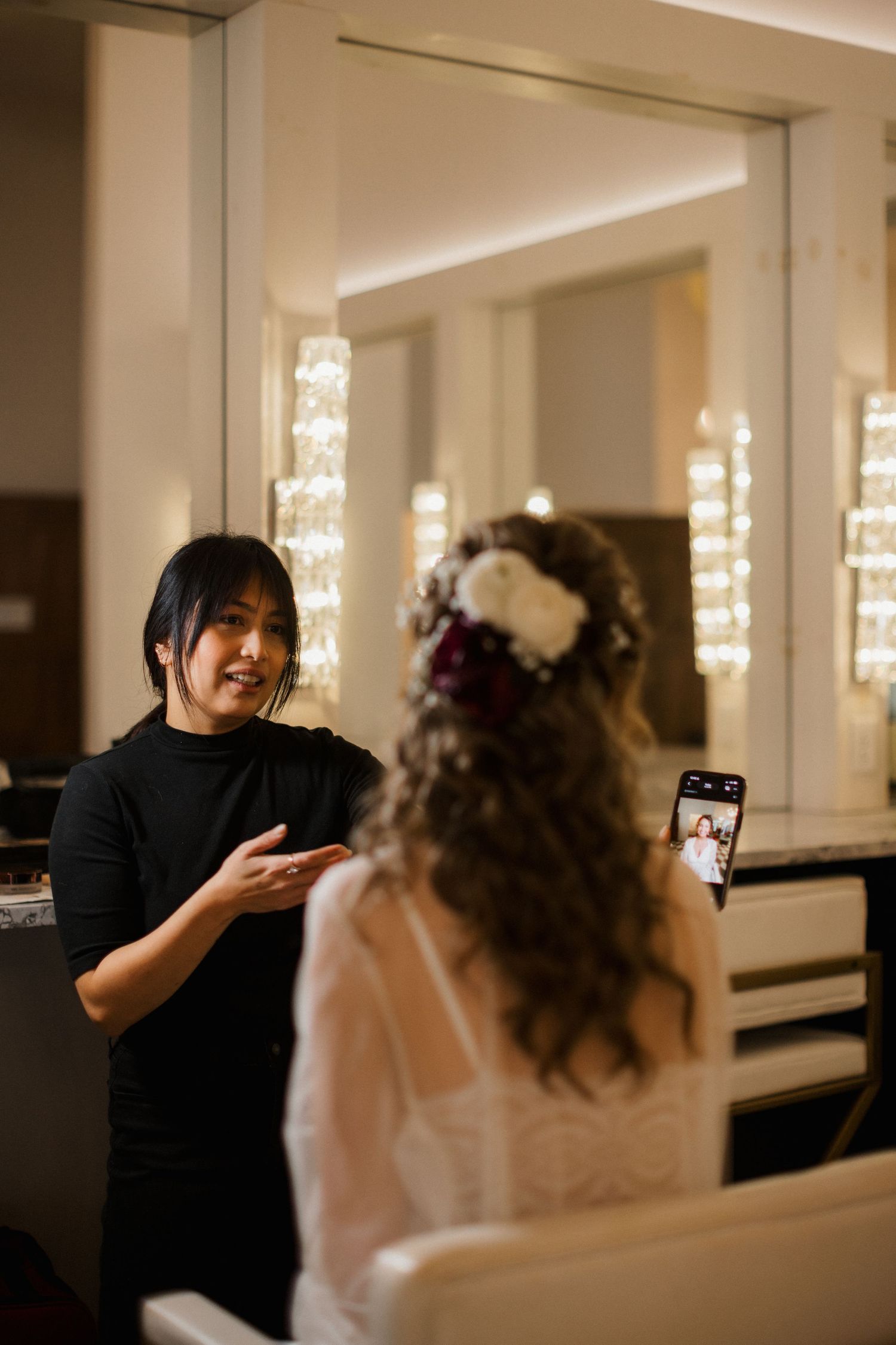 Makeup artist talking with a bride in a lace gown and floral hair piece at a glamorous salon mirror before the wedding.