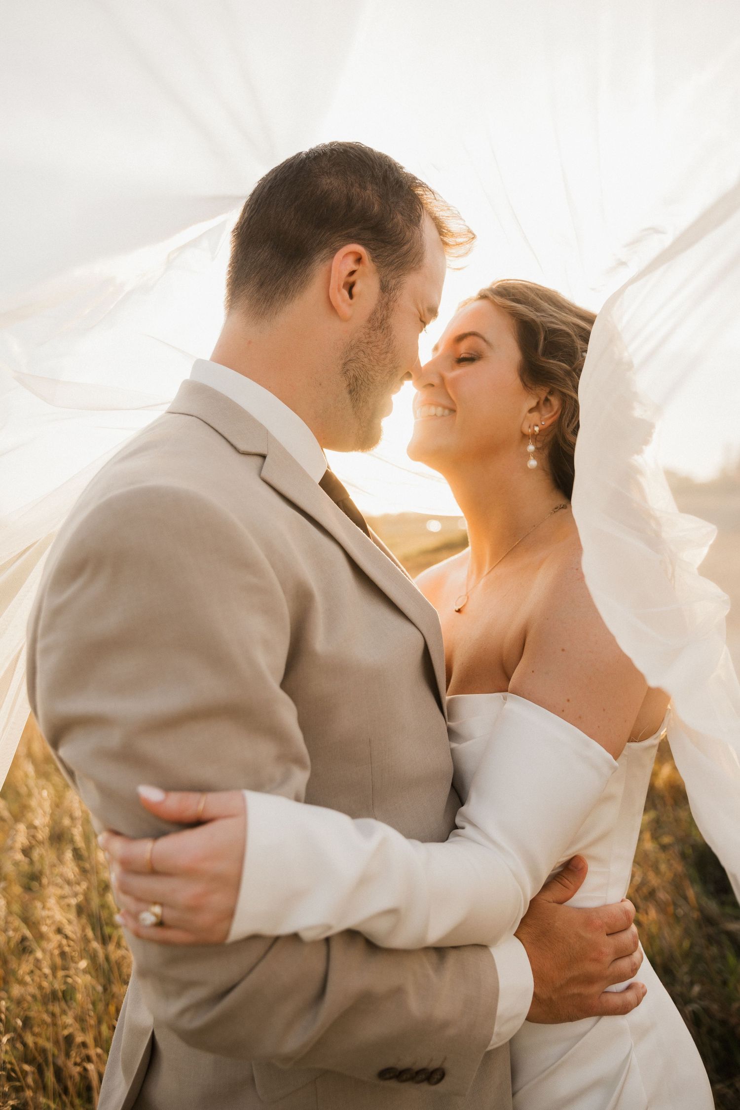 Smiling bride with closed eyes nuzzles her bearded groom in a tan suit under a flowing veil in a golden field.