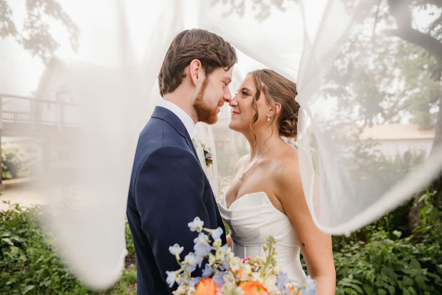 Bride and groom share a romantic moment under a flowing veil, surrounded by lush greenery on their wedding day.