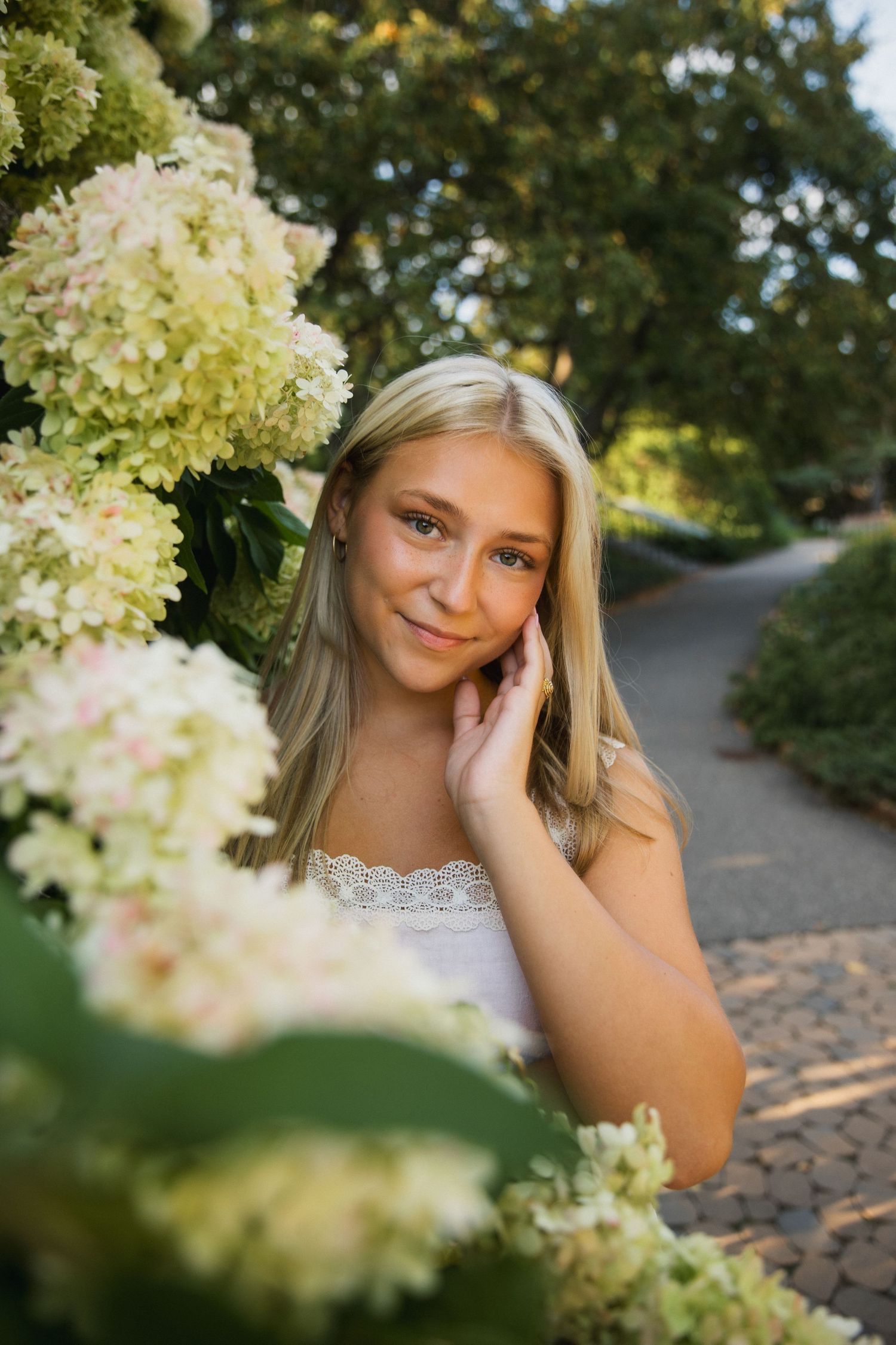 Young blonde woman posing gracefully among white hydrangea blooms in a sunlit garden path, wearing a white lace top.