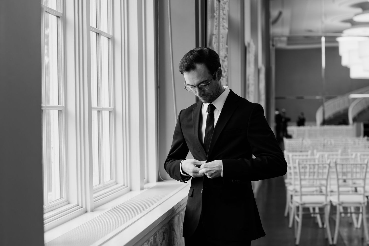 Black and white photo of a groom in a suit adjusting his cufflinks by large windows before his wedding ceremony.