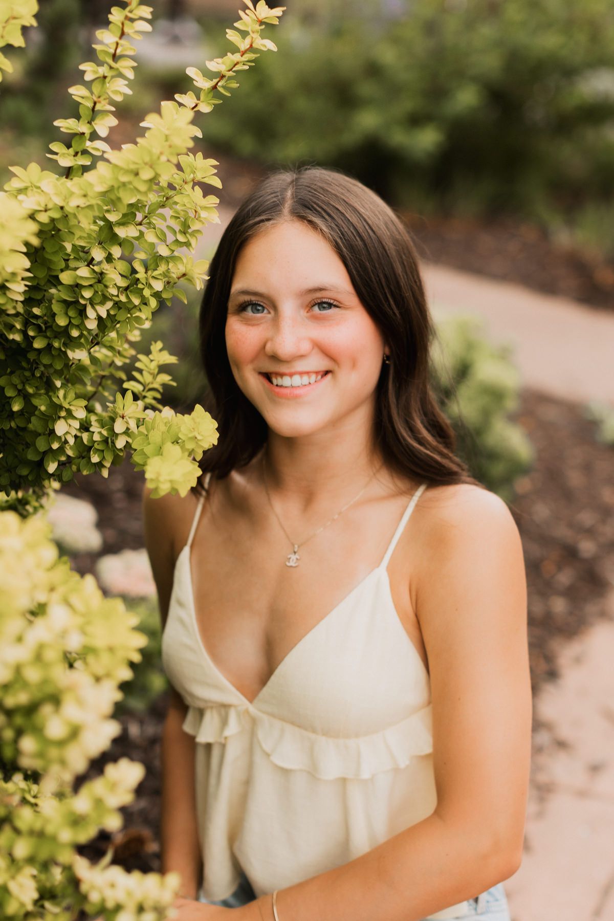 Smiling brunette teen girl in cream camisole posing beside bright green garden shrubs.