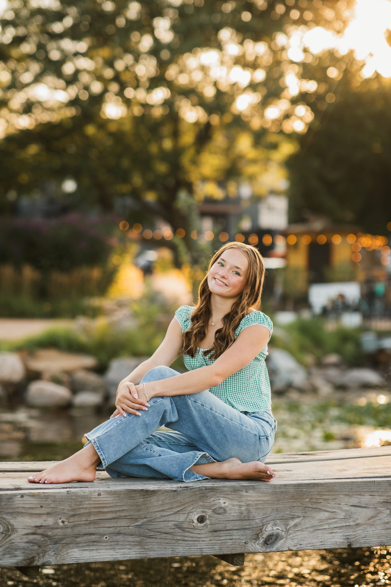 Young woman in green top and jeans sitting barefoot on wooden dock, smiling at golden hour sunset.