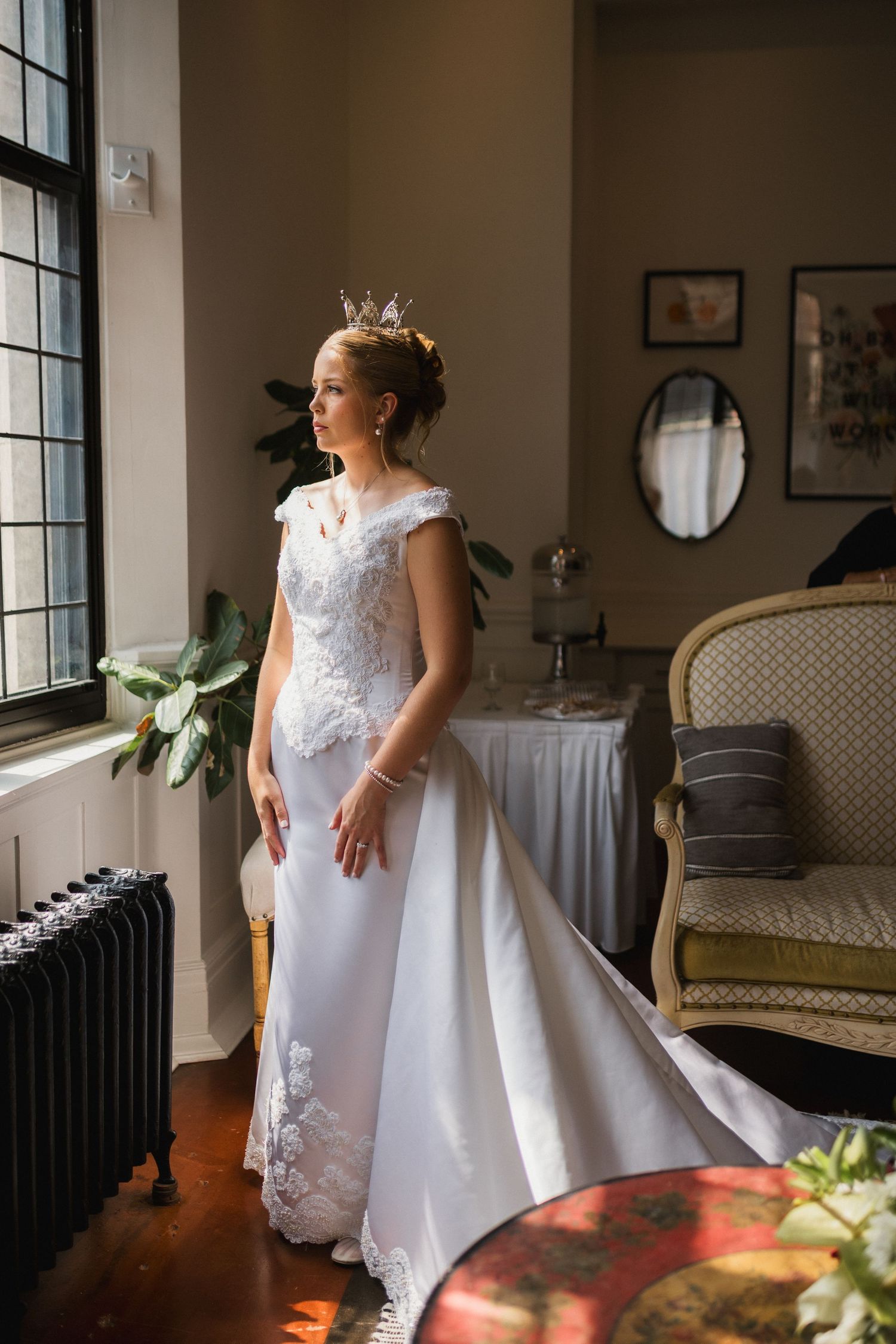Elegant bride wearing a crown and lace wedding gown standing by a window in a vintage room.
