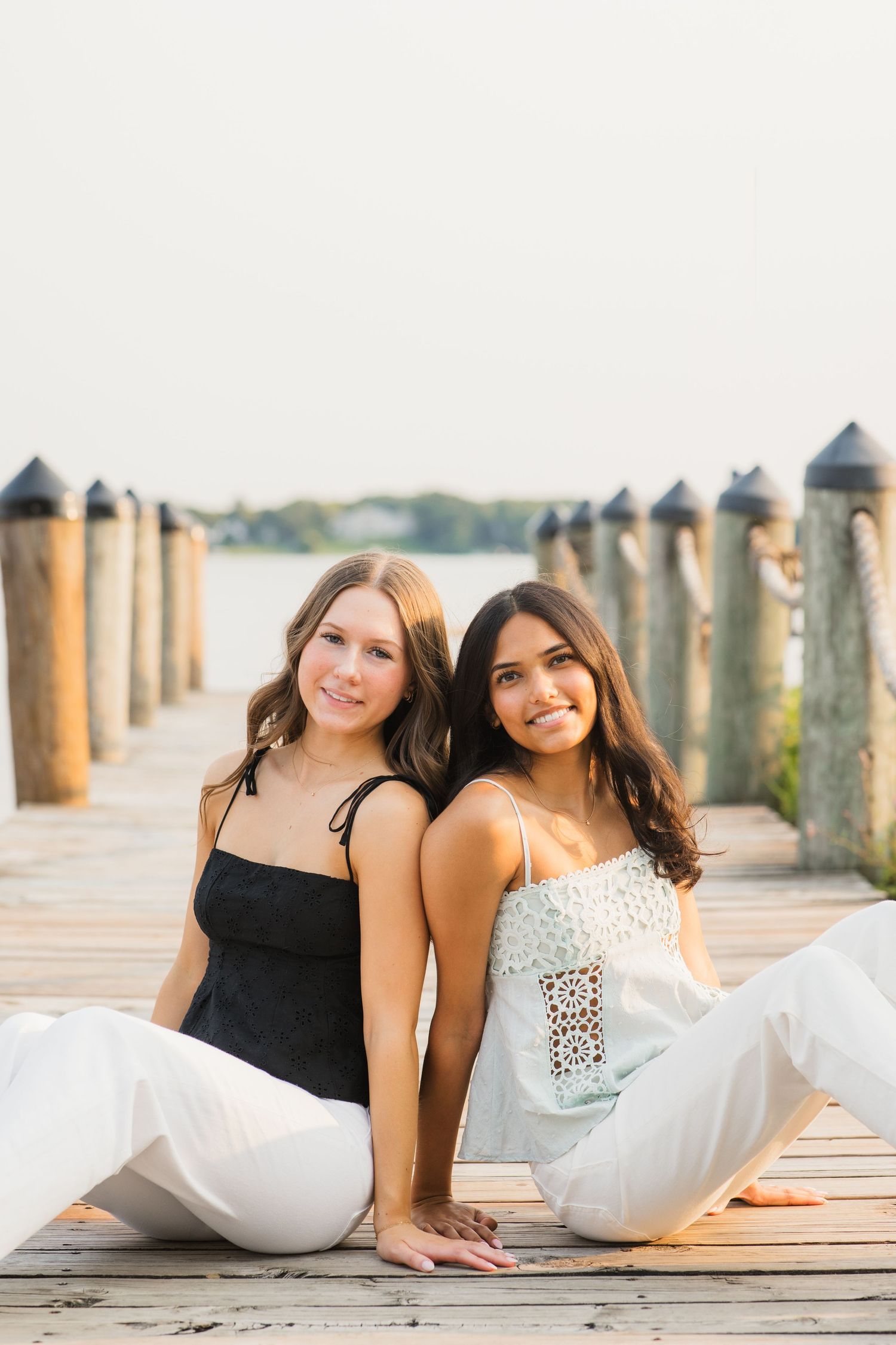 Two young women sitting back-to-back on a wooden dock near water, wearing white pants and smiling at the camera.