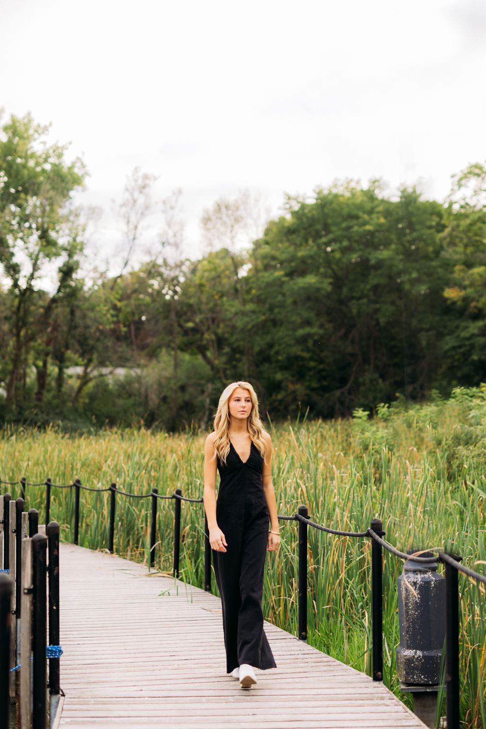Woman in long black dress stands on wooden boardwalk through natural wetland area on sunny summer day.