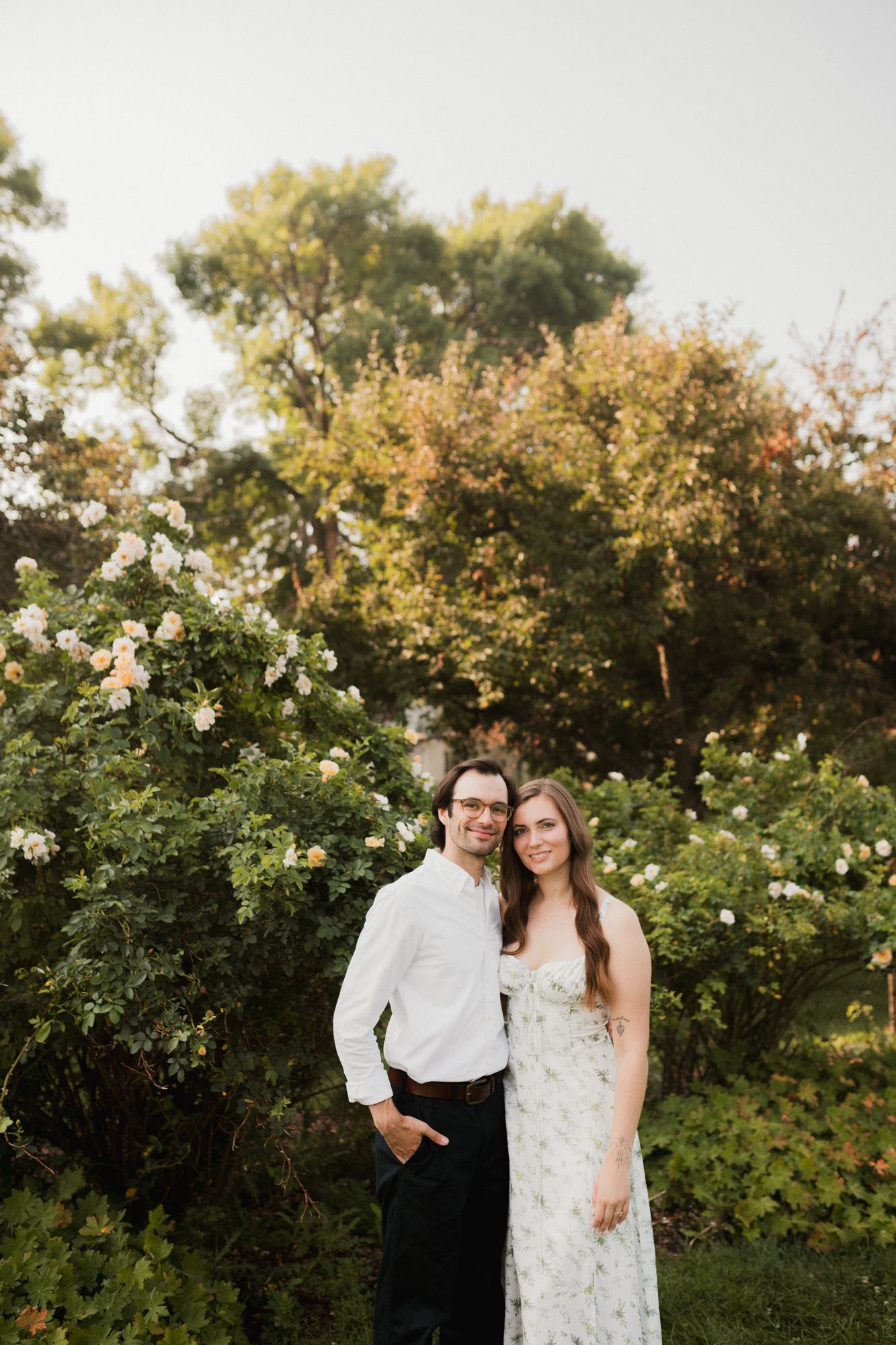 Smiling couple posing together in a garden with blooming white roses and trees in soft light.