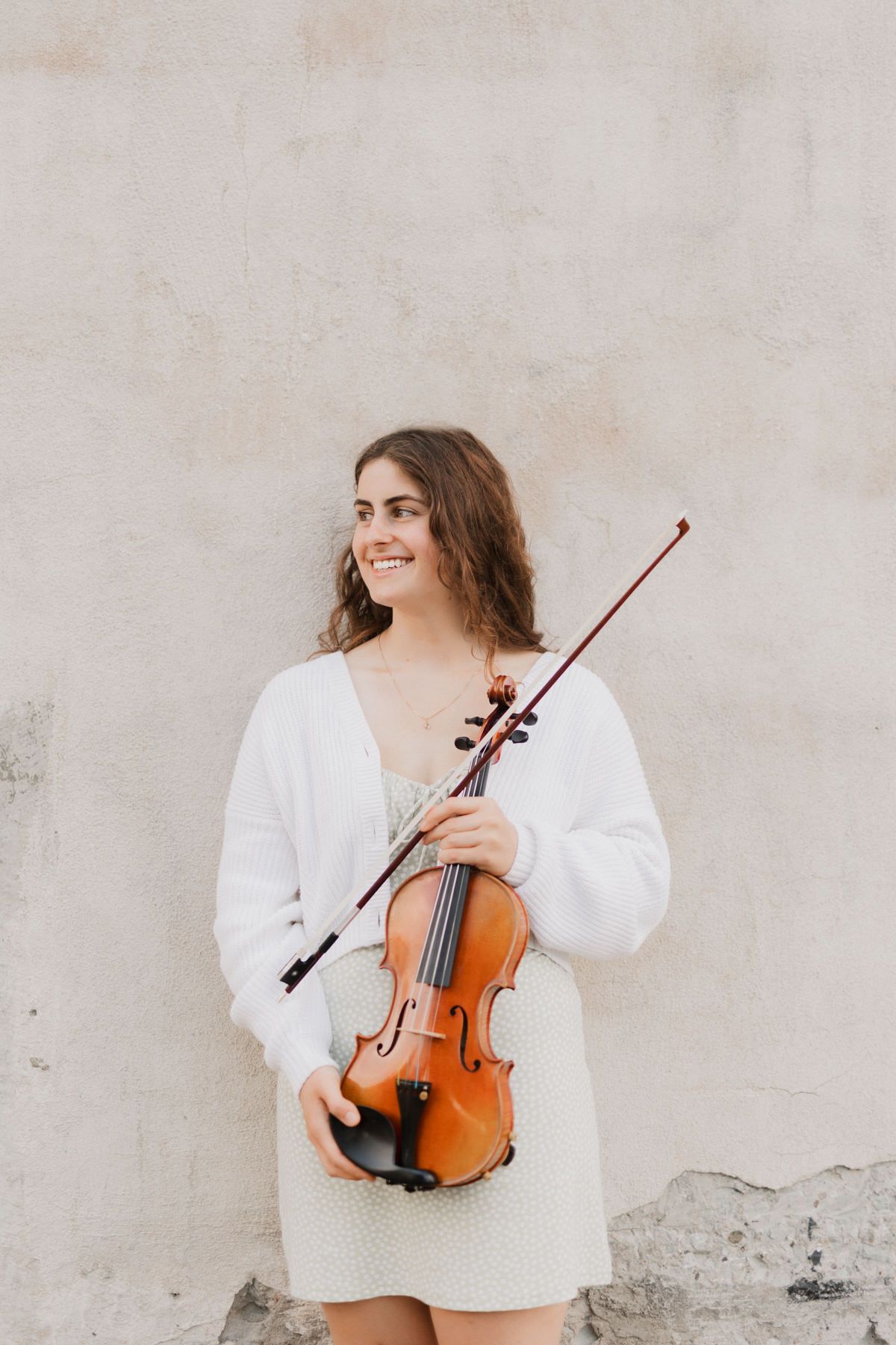 Smiling young woman holding a violin and bow against a neutral wall, wearing a white cardigan and dress.