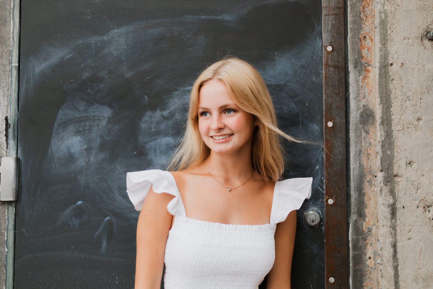 Smiling blonde woman in white ruffle dress poses against rustic dark metal door outdoors.