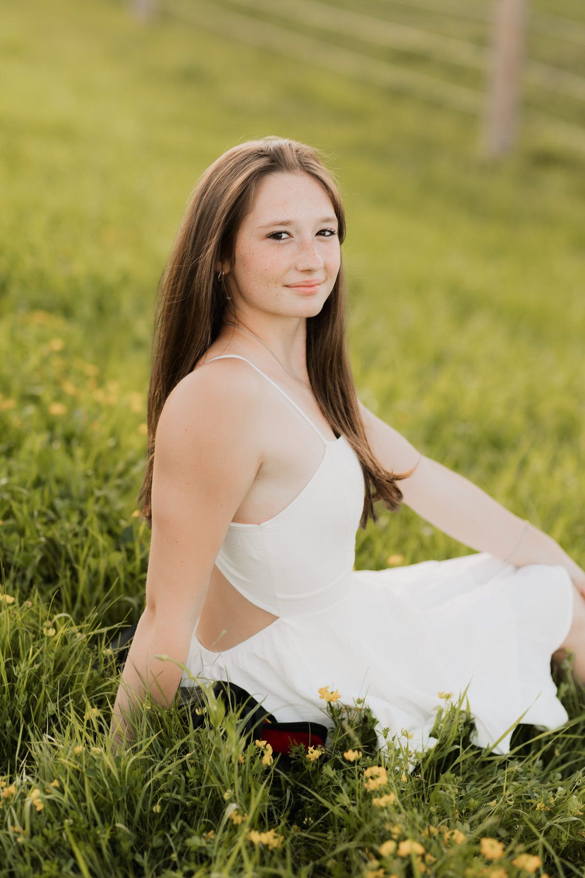 Young woman with long brown hair in white dress sitting in sunny wildflower meadow.