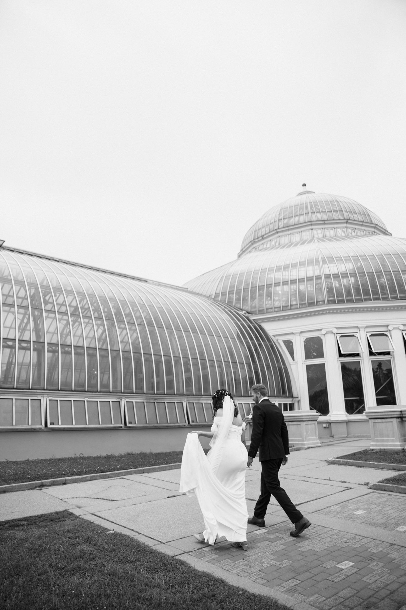 Black and white photo of a bride and groom walking hand in hand outside a grand greenhouse conservatory.