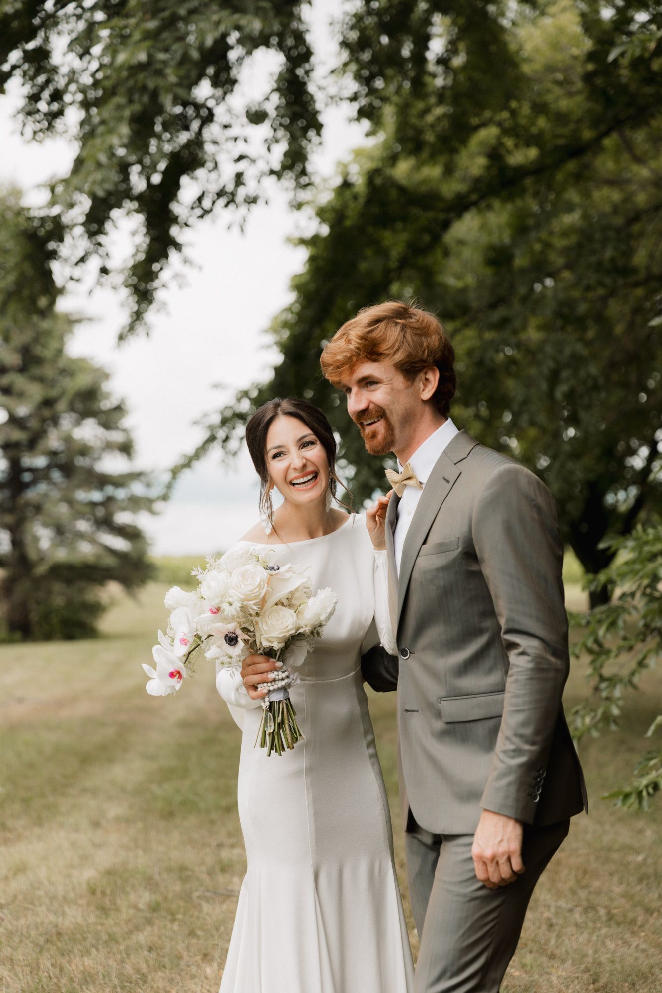 Smiling bride in white gown holding floral bouquet stands with groom in gray suit outdoors under lush green trees.