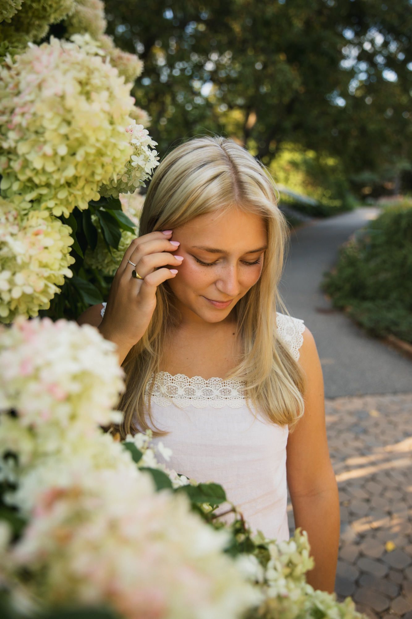 Young blonde woman in white top smelling hydrangea flowers in a sunlit garden path.