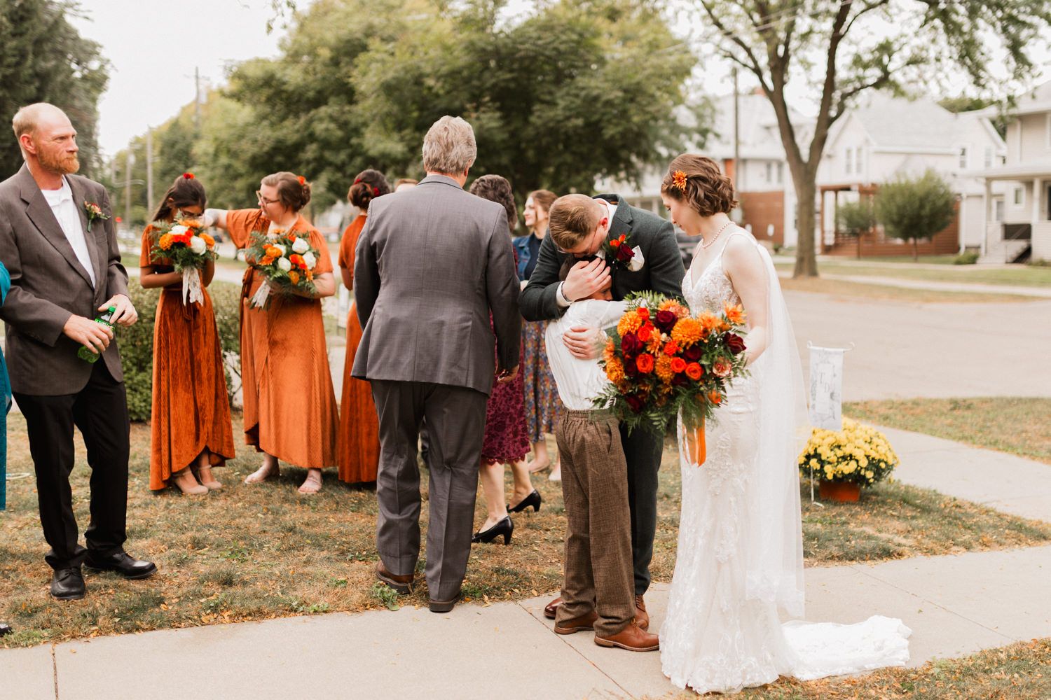 Wedding party gathered outdoors with orange bridesmaid dresses and bright floral arrangements.