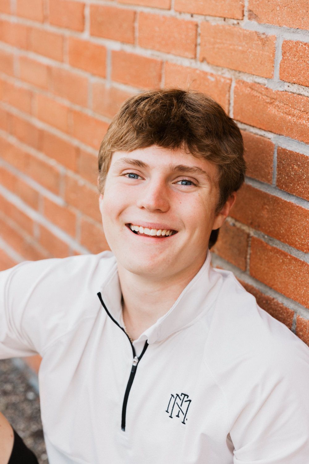 Subject in a white quarter-zip pullover with embroidered logo posing against a brick wall in a casual portrait.