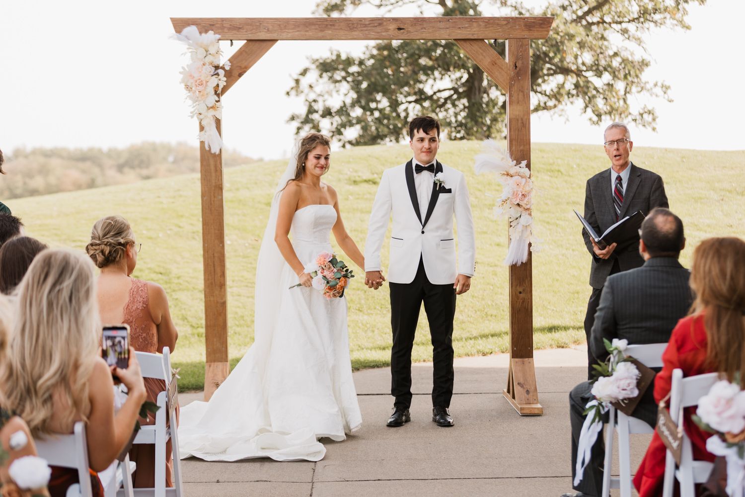 A wedding ceremony takes place under a rustic wooden pavilion in an outdoor setting during golden hour.