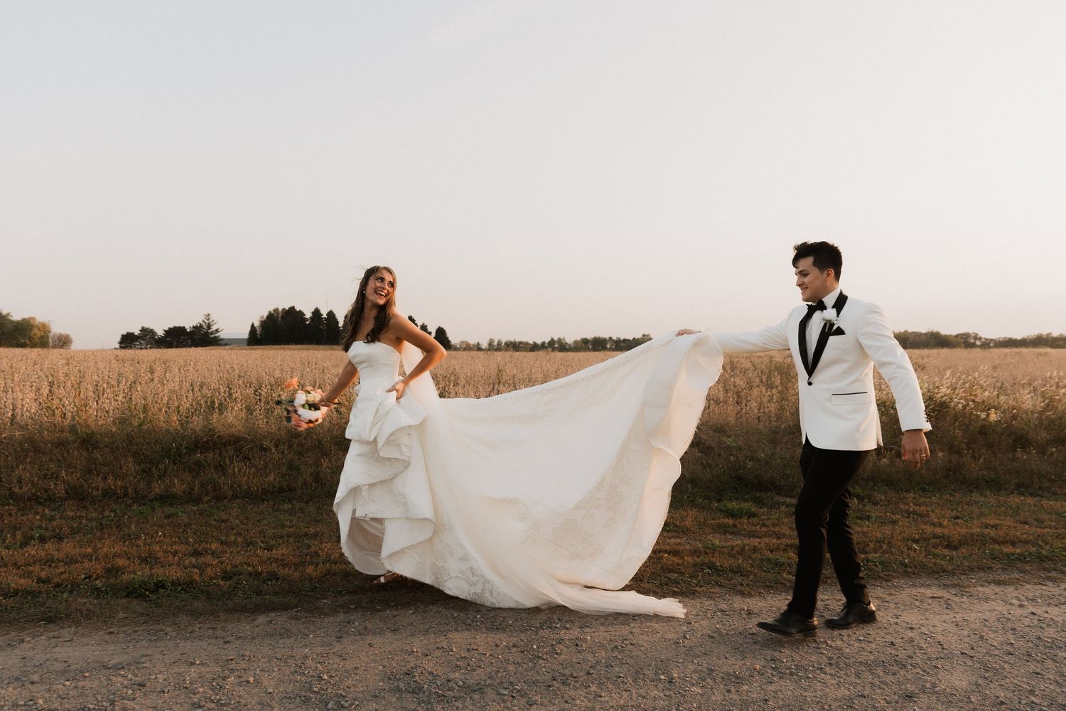 Bride in white gown and groom in white tuxedo walking on a rural path with golden fields at sunset.