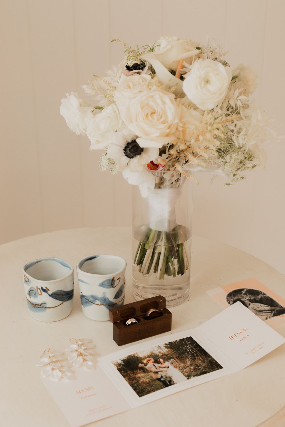 White floral bouquet in vase with matching ceramic mugs, photos and accessories arranged on white table in warm lighting.