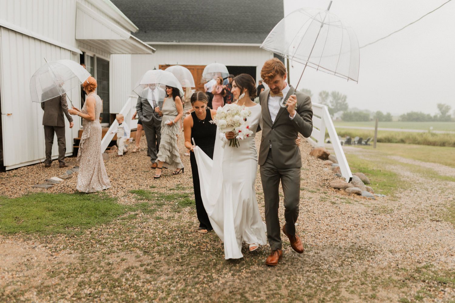Wedding party members walk and pose along a gravel path outside on an overcast day.