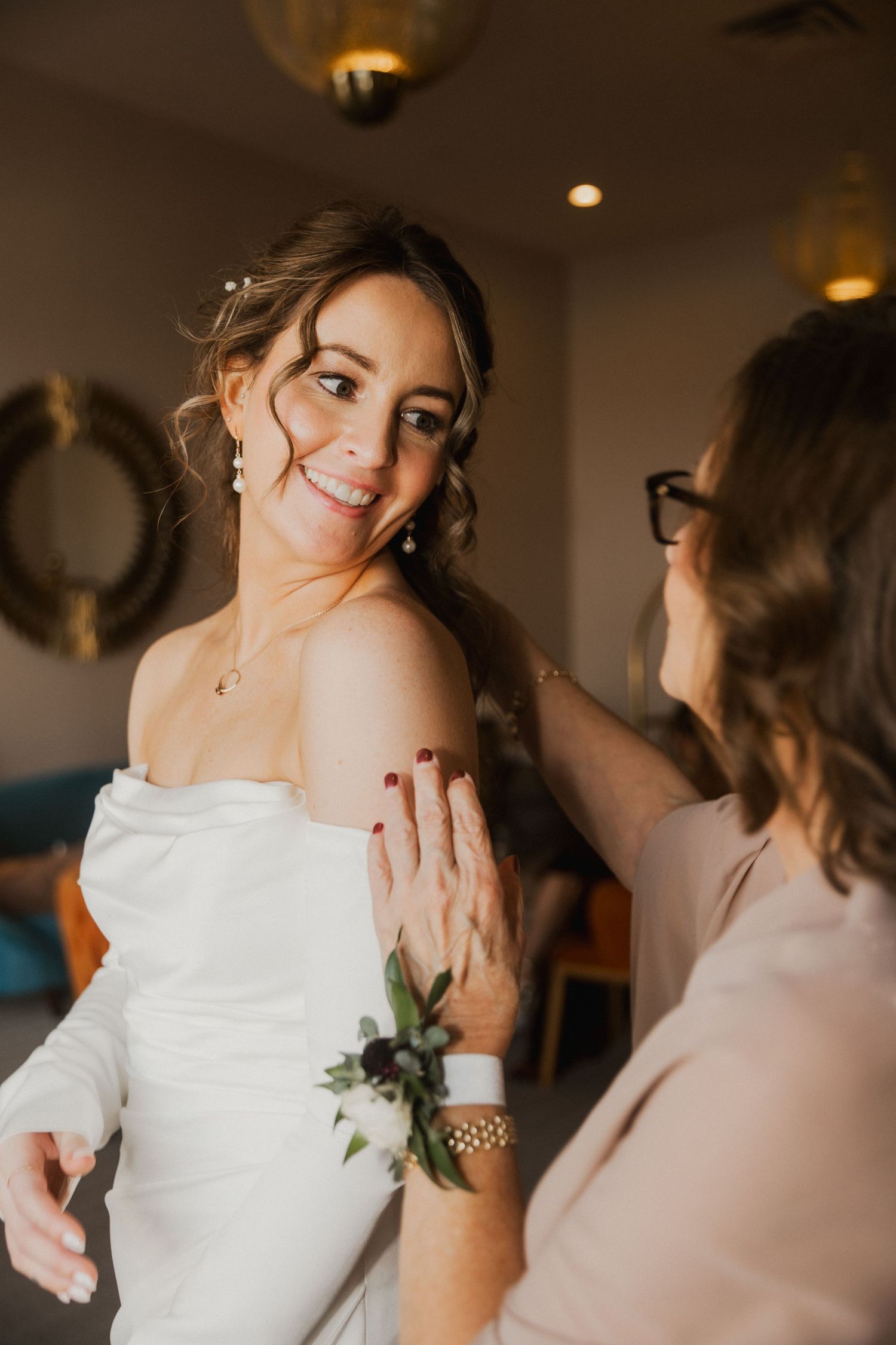 Smiling bride in a white off-the-shoulder gown with a floral wrist corsage is helped by a woman in glasses.
