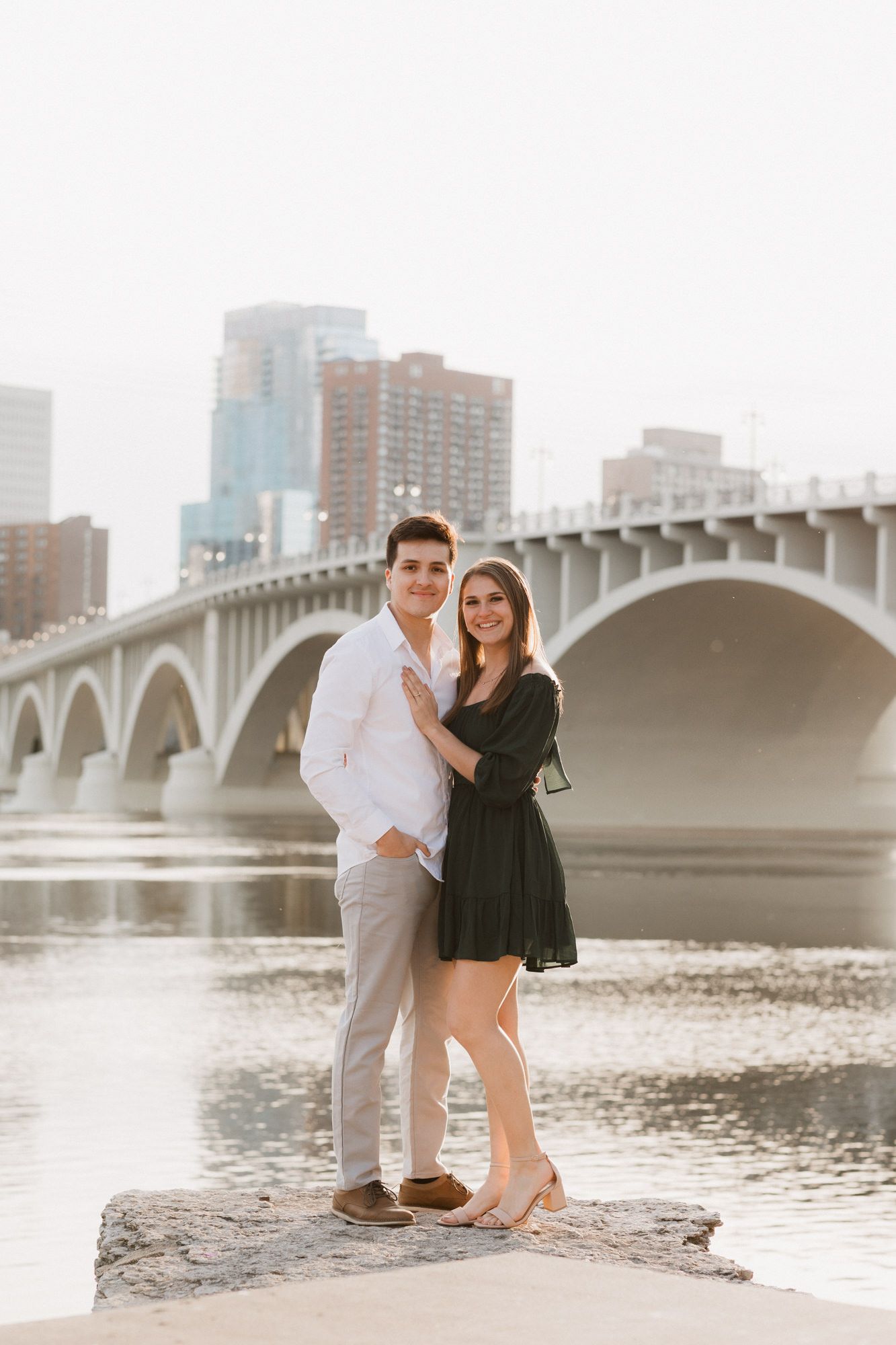 Couple posing on rocks by a river with a bridge and city skyline in the background.