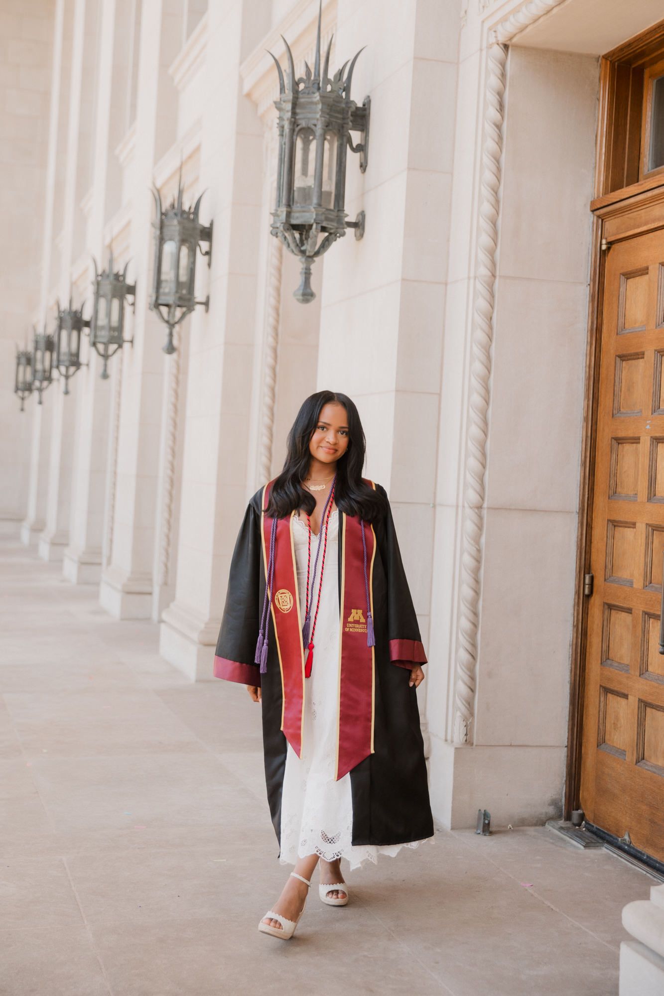 Young woman in black graduation gown with maroon stole walks along elegant columned building exterior.