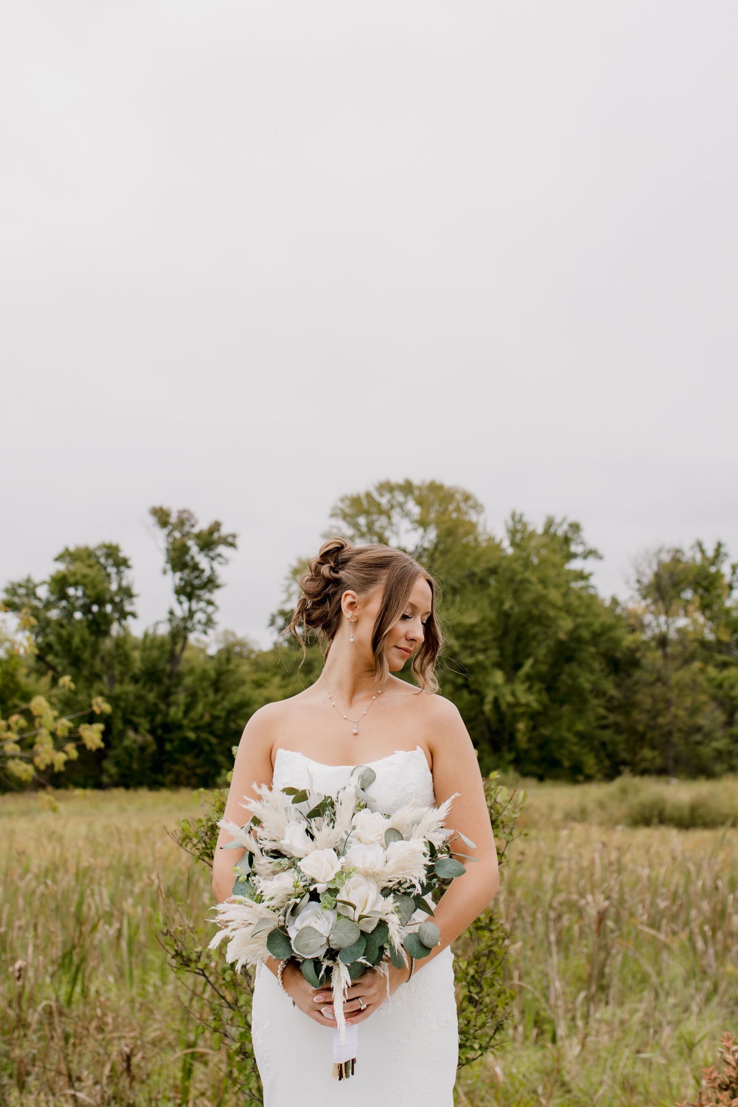 Bride in white strapless gown holding white floral bouquet with greenery, standing in a field with trees in background.