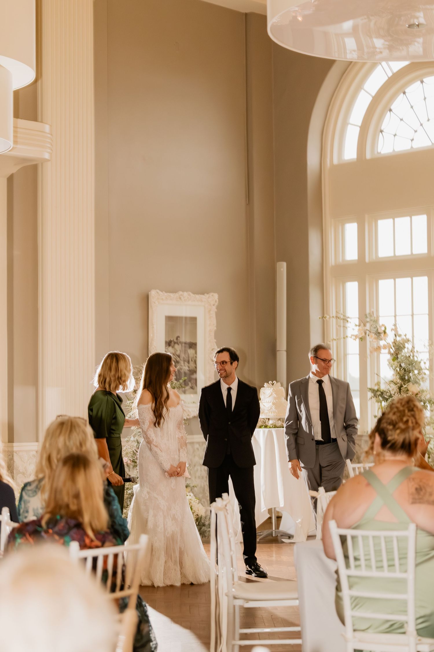 Elegant wedding ceremony in a bright ballroom with bride and groom at the altar, guests seated in white chairs.