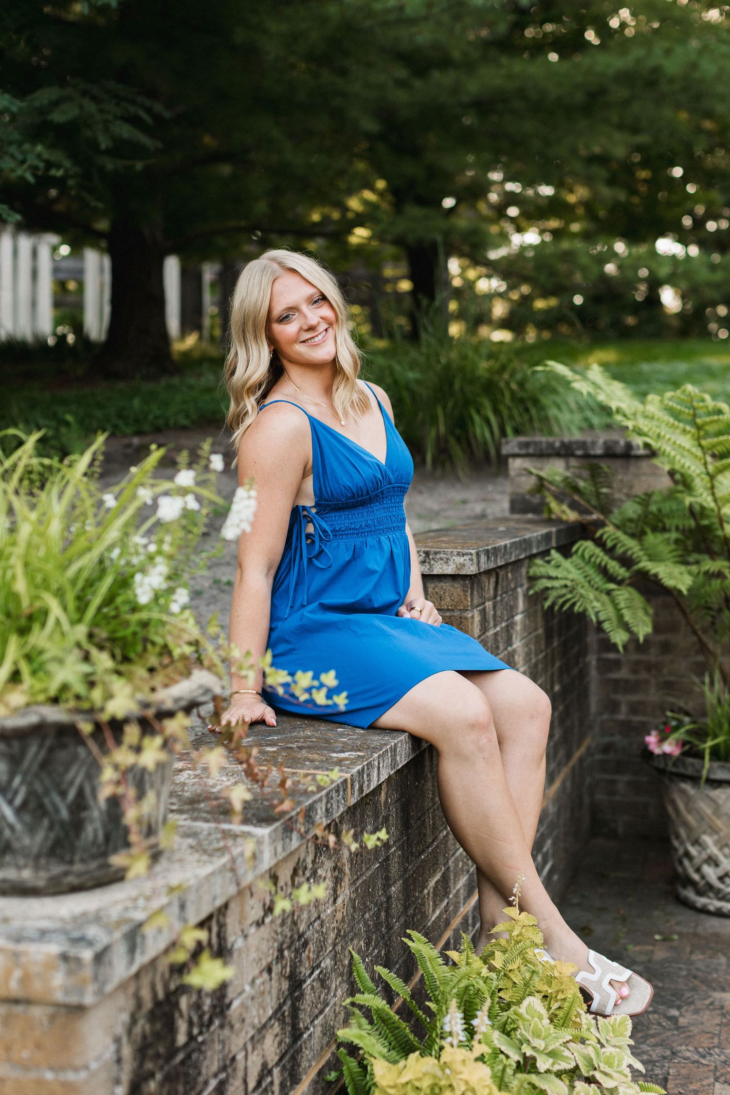 Person in blue dress sitting on stone garden wall surrounded by lush ferns and summer foliage.