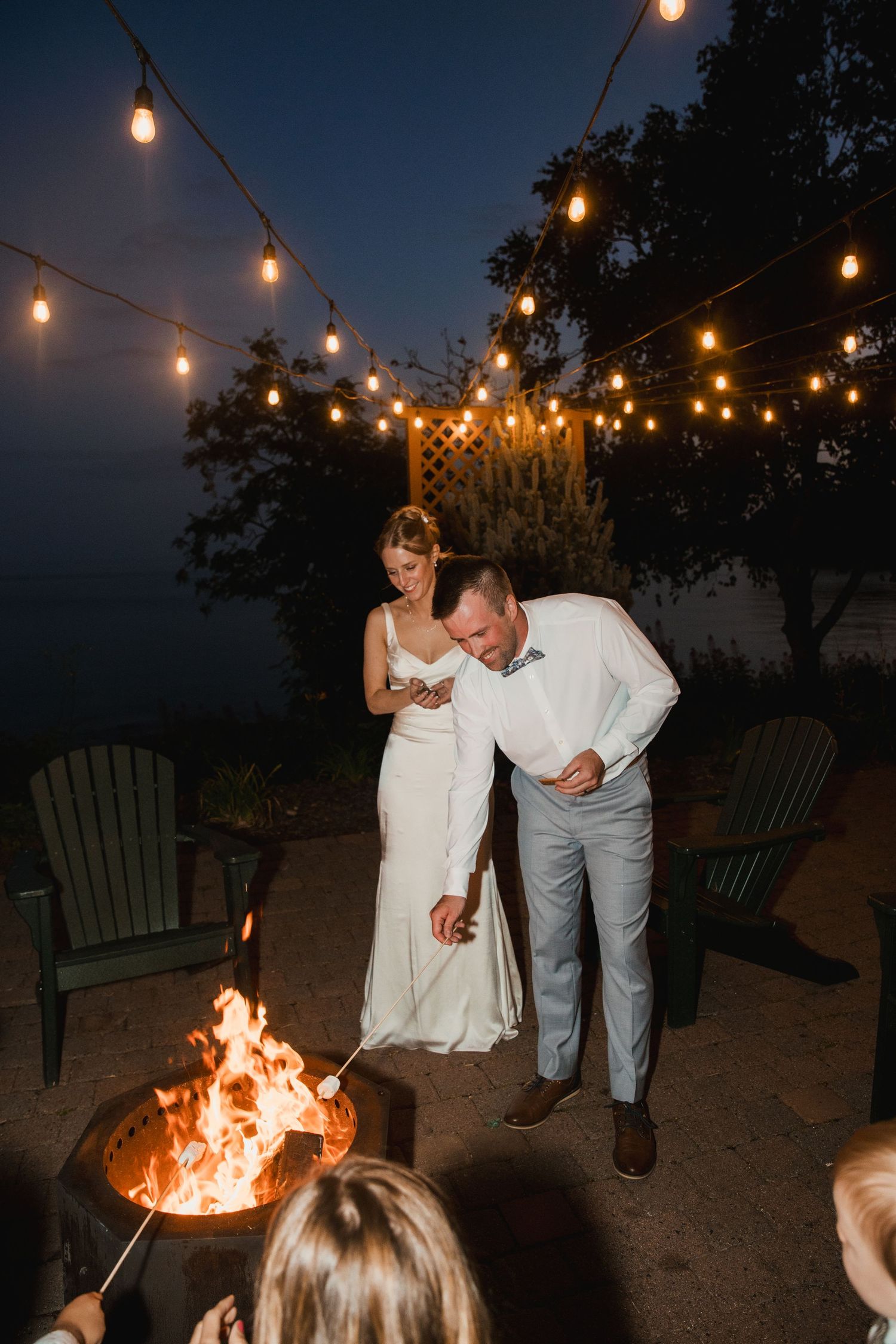 Bride and groom laughing by a fire pit at night, surrounded by string lights at an outdoor wedding reception.